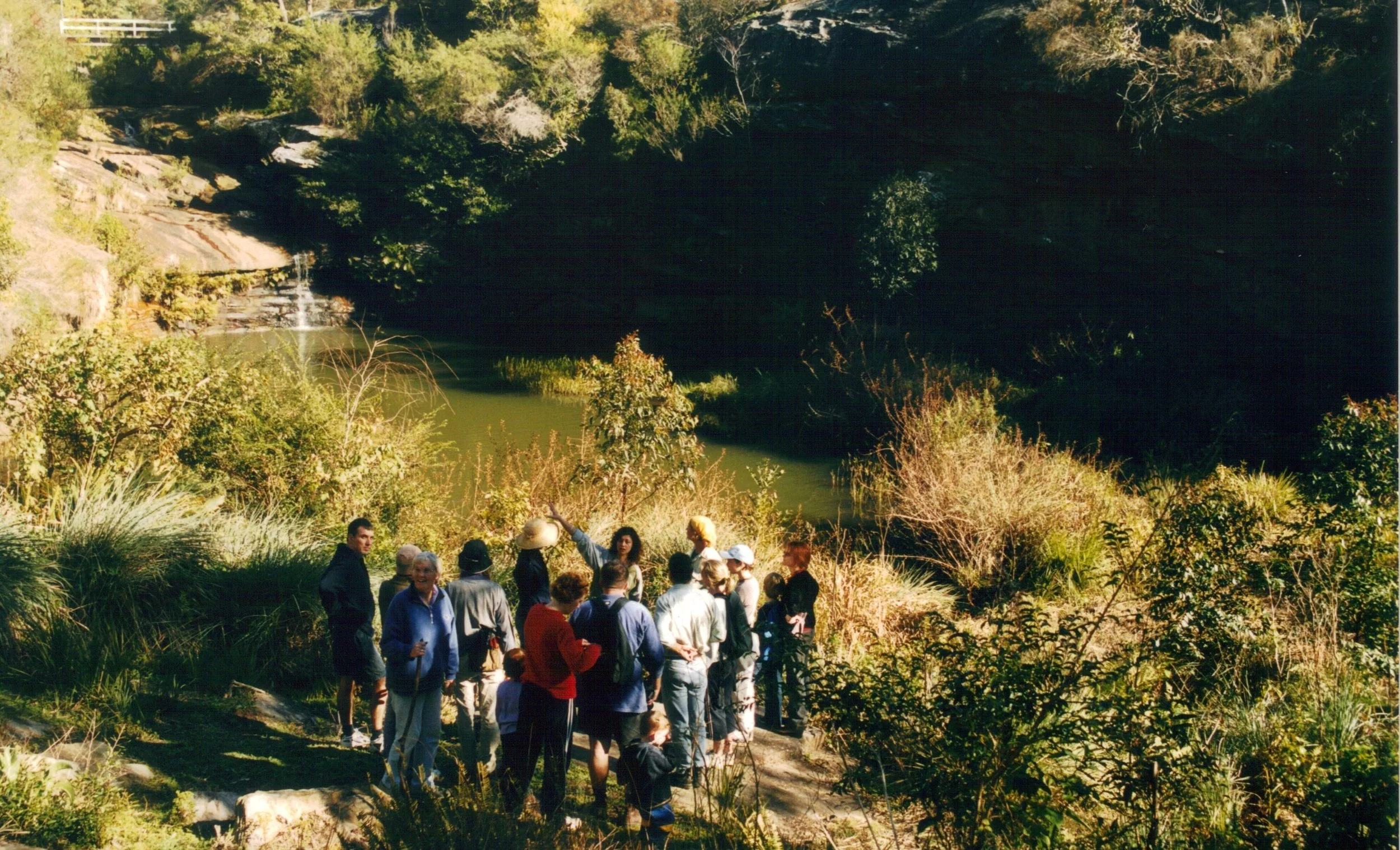 A group of people gathered outdoors near a river and lush greenery, possibly on a guided nature tour or educational outing.