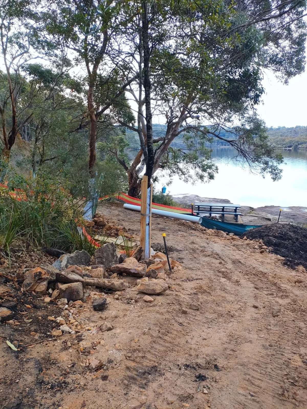 A dirt pathway leading to a body of water, with a small rock pile and a tree in the foreground. There is construction fencing and pipes on the dirt path, with trees and water in the background.