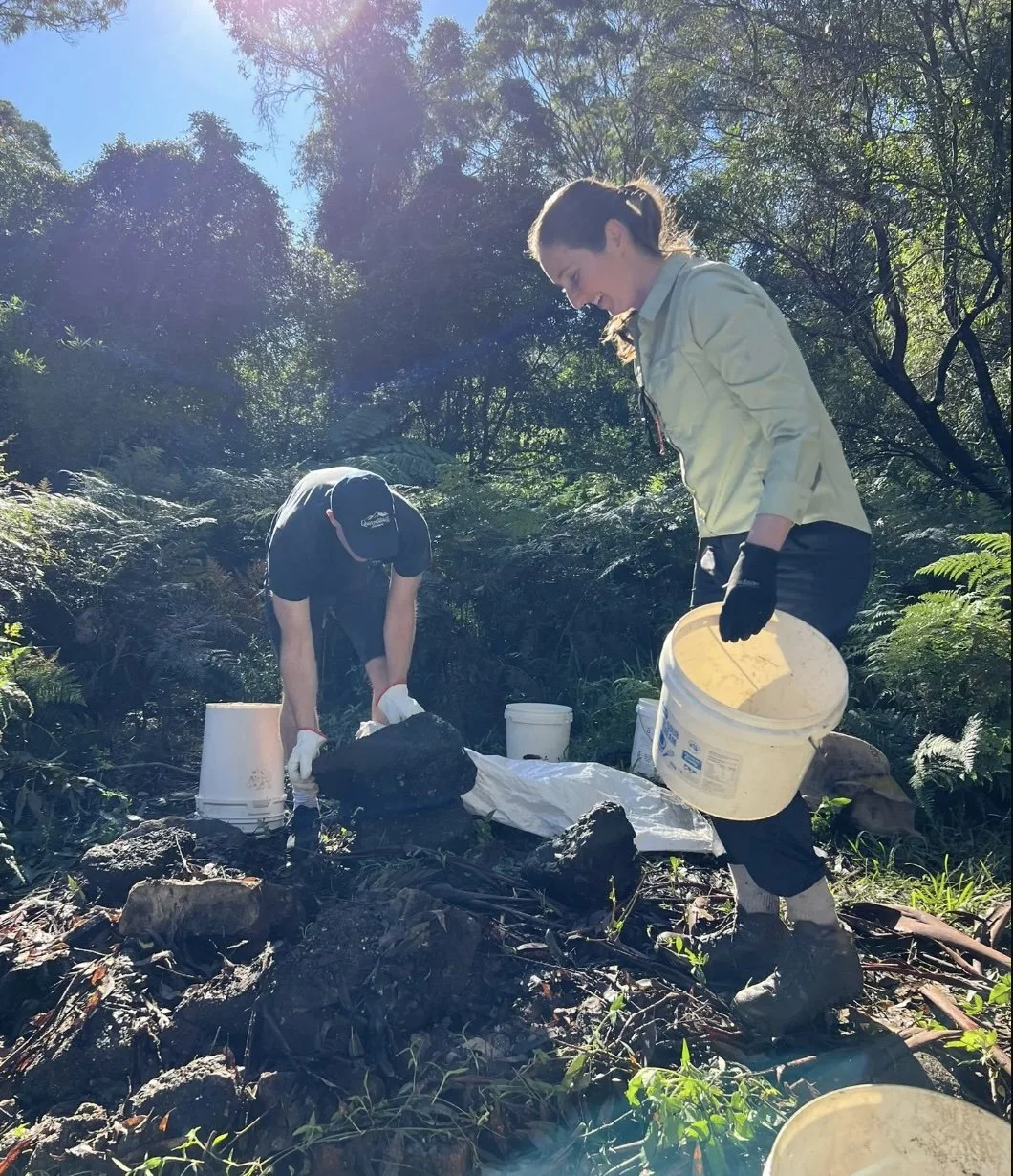 Two women working outdoors in a forest, handling soil and tools, with buckets and a white cloth on the ground, sunlight filtering through trees.