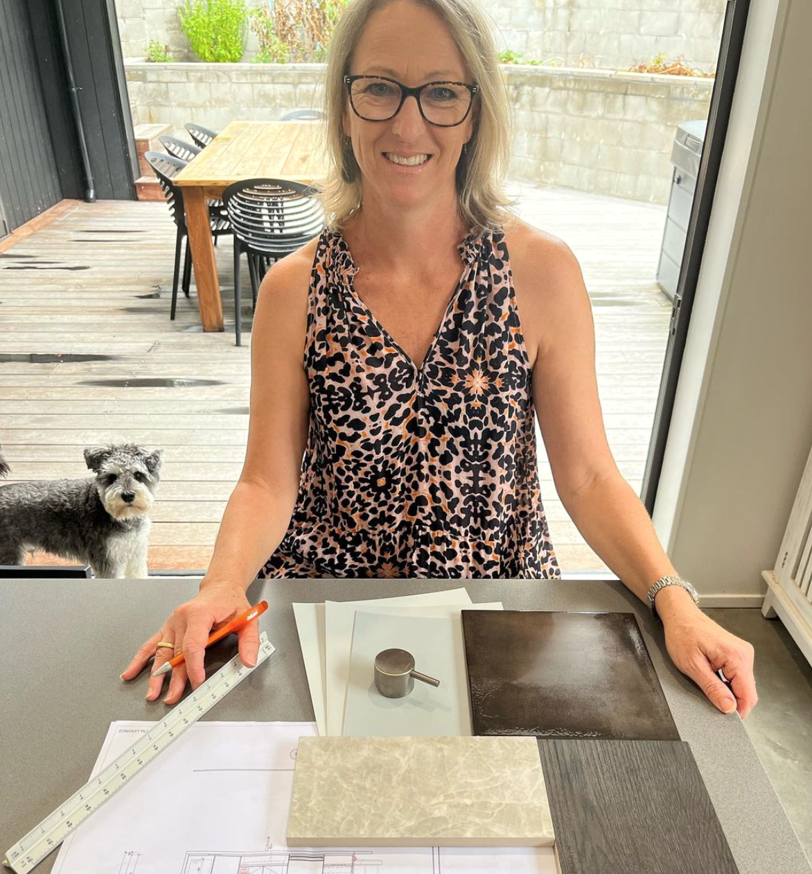 A woman with blonde hair, glasses, and a leopard-print sleeveless dress sitting at a table with architectural plans, samples, and a metal cup, smiling at the camera. Outside, a small dog stands on a wooden deck near a table and chairs.