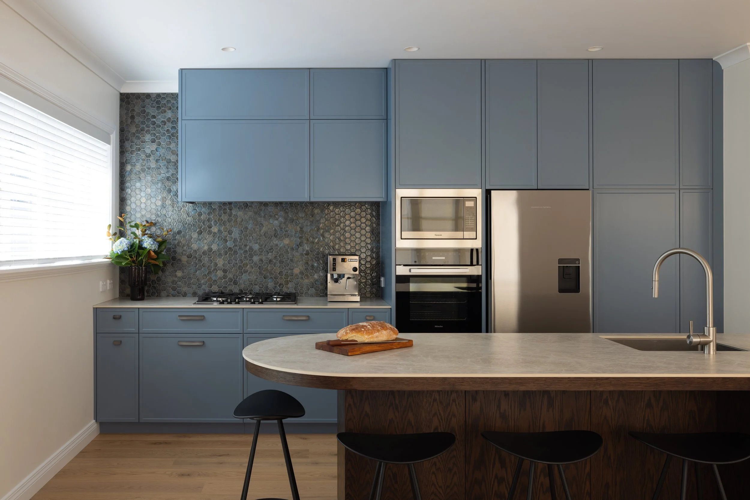 Modern kitchen with blue cabinets, a marble island, and stainless steel appliances, including a refrigerator and microwave. There is a loaf of bread on the island and a flower vase near the window.