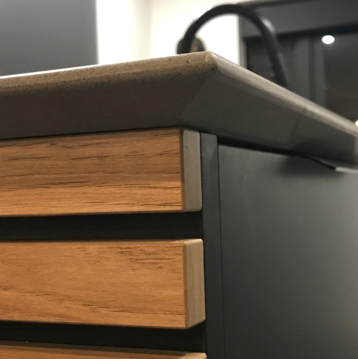 Close-up of a modern desk with wooden drawers and a black frame, with a grey countertop. Part of a printer or computer is visible in the background.
