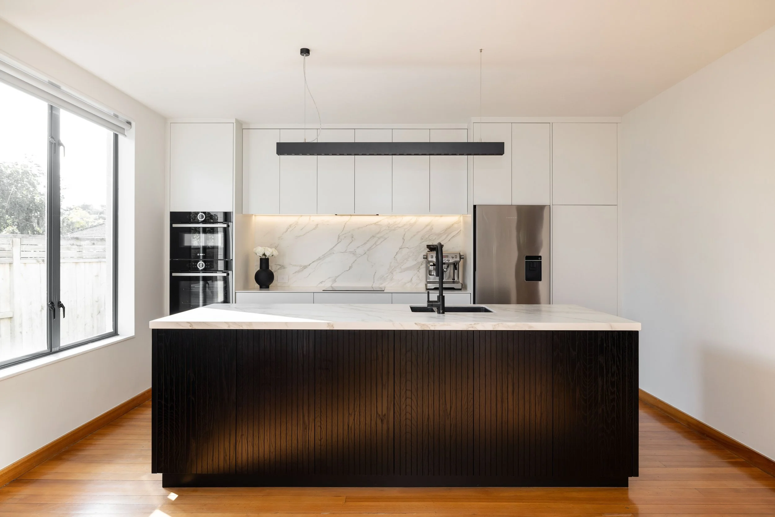Modern kitchen with a black kitchen island, white cabinetry, marble backsplash, stainless steel refrigerator, built-in oven, and a coffee machine, with hardwood floors and large window.