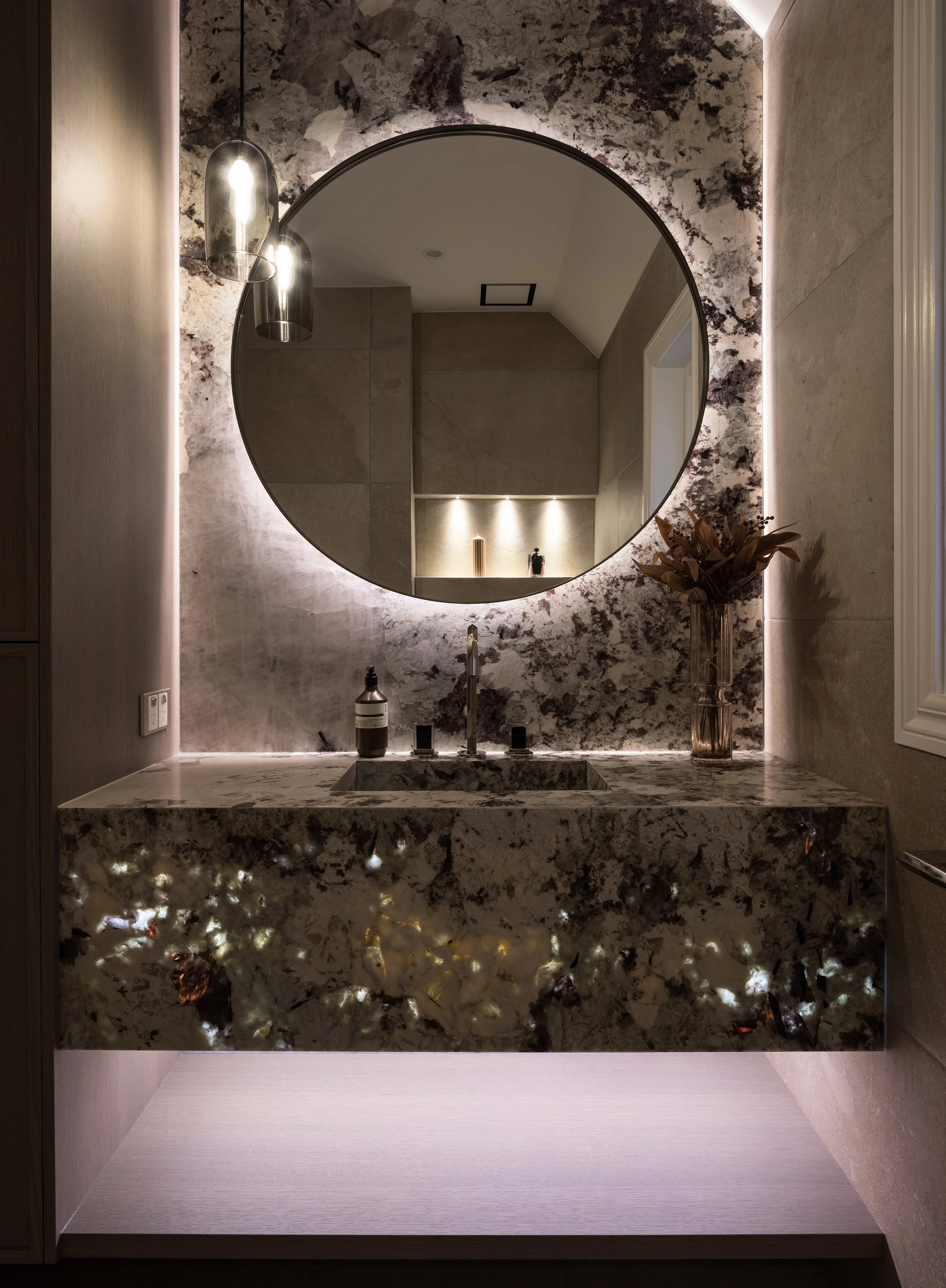 A modern bathroom with a large round mirror, backlit with LED lights, above a Speckled granite vanity with a built-in sink. To the right, a tall vase with dried flowers sits on the counter. On the left, three metallic pendant lights hang from the ceiling.