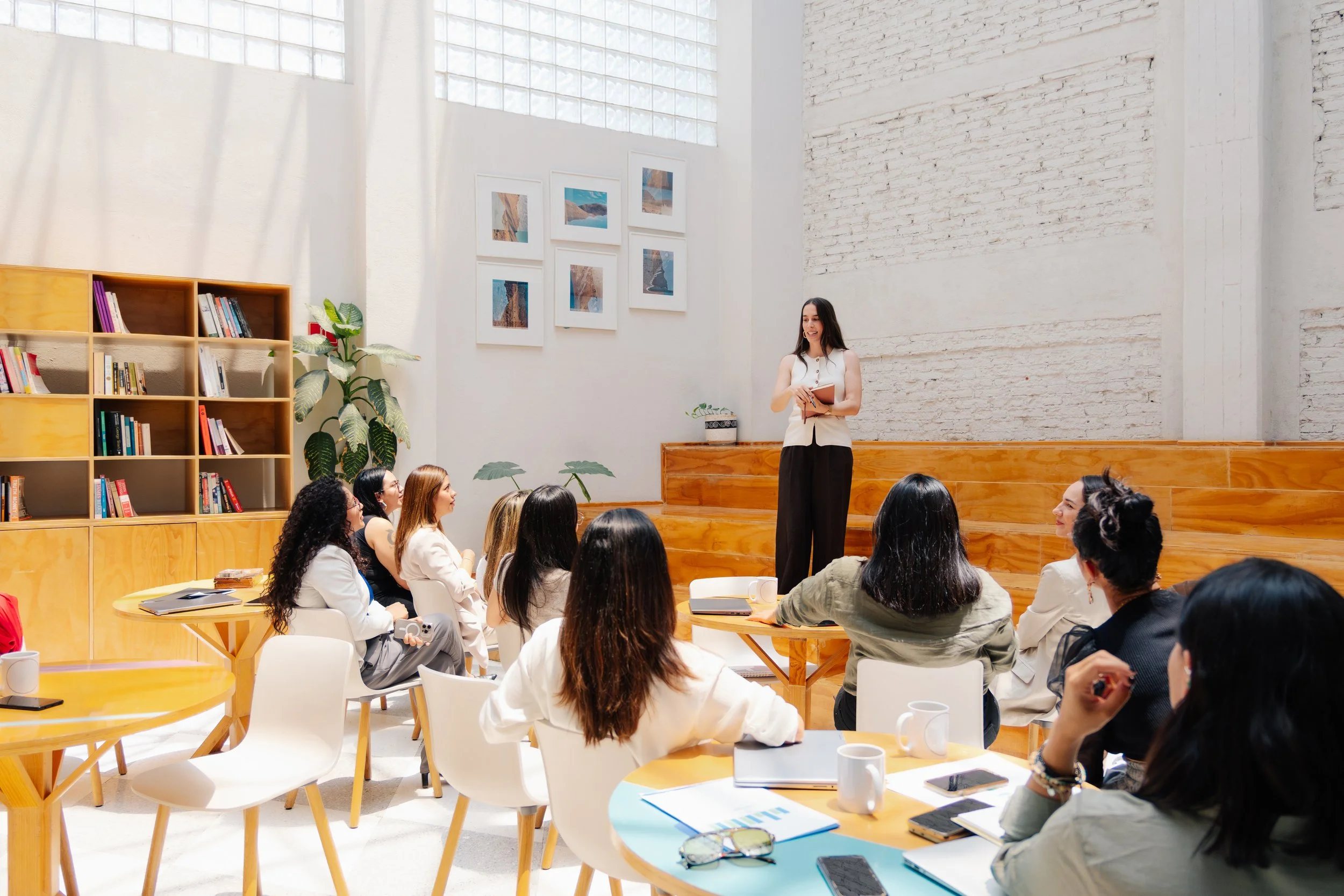 Una mujer da una presentación a un grupo de personas en una sala luminosa con paredes de ladrillo blanco, estanterías de madera y cuadros colgados.