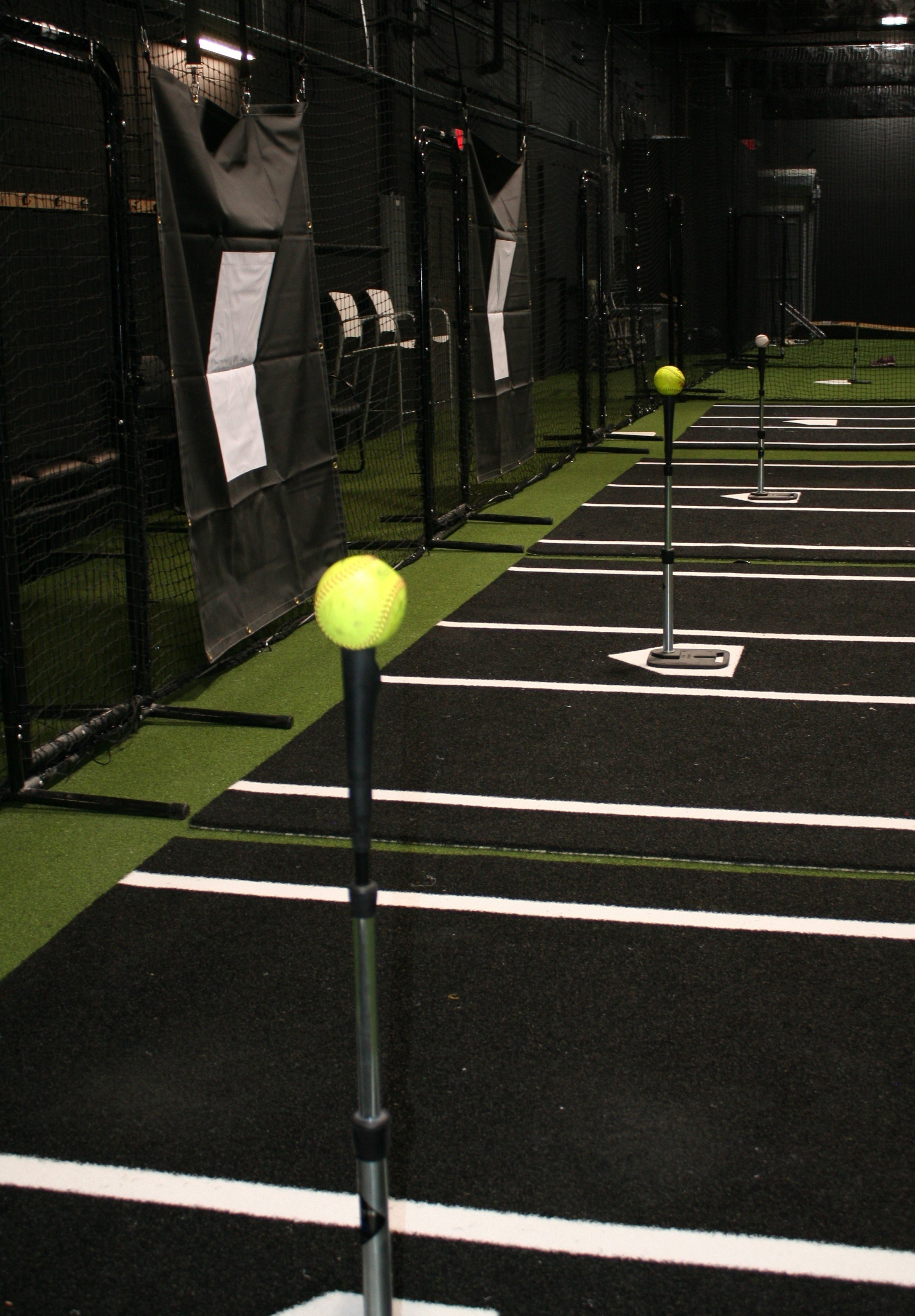 Indoor baseball or softball training facility with pitching lanes, each marked with a number, and yellow softballs on pitching machines or stands.