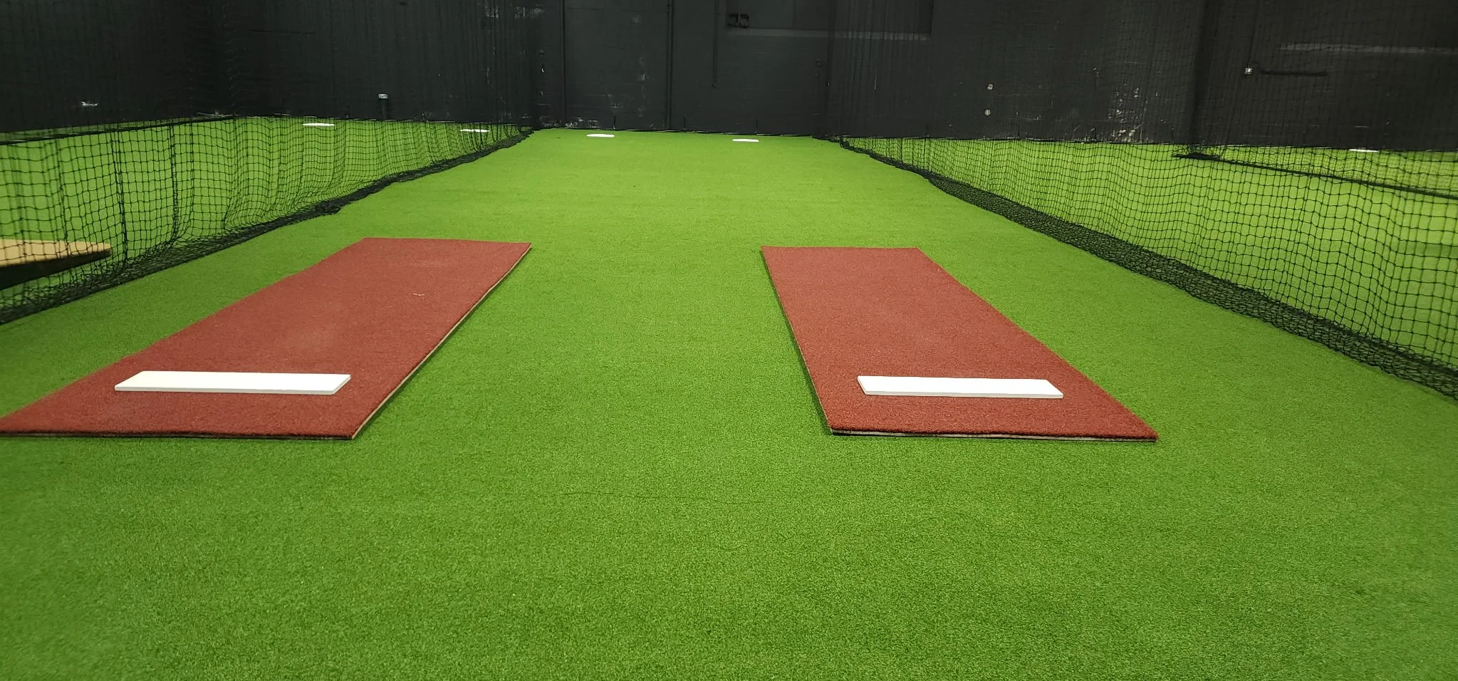 Indoor pitching practice area with artificial green turf, two red mats, and white pitching rubbers