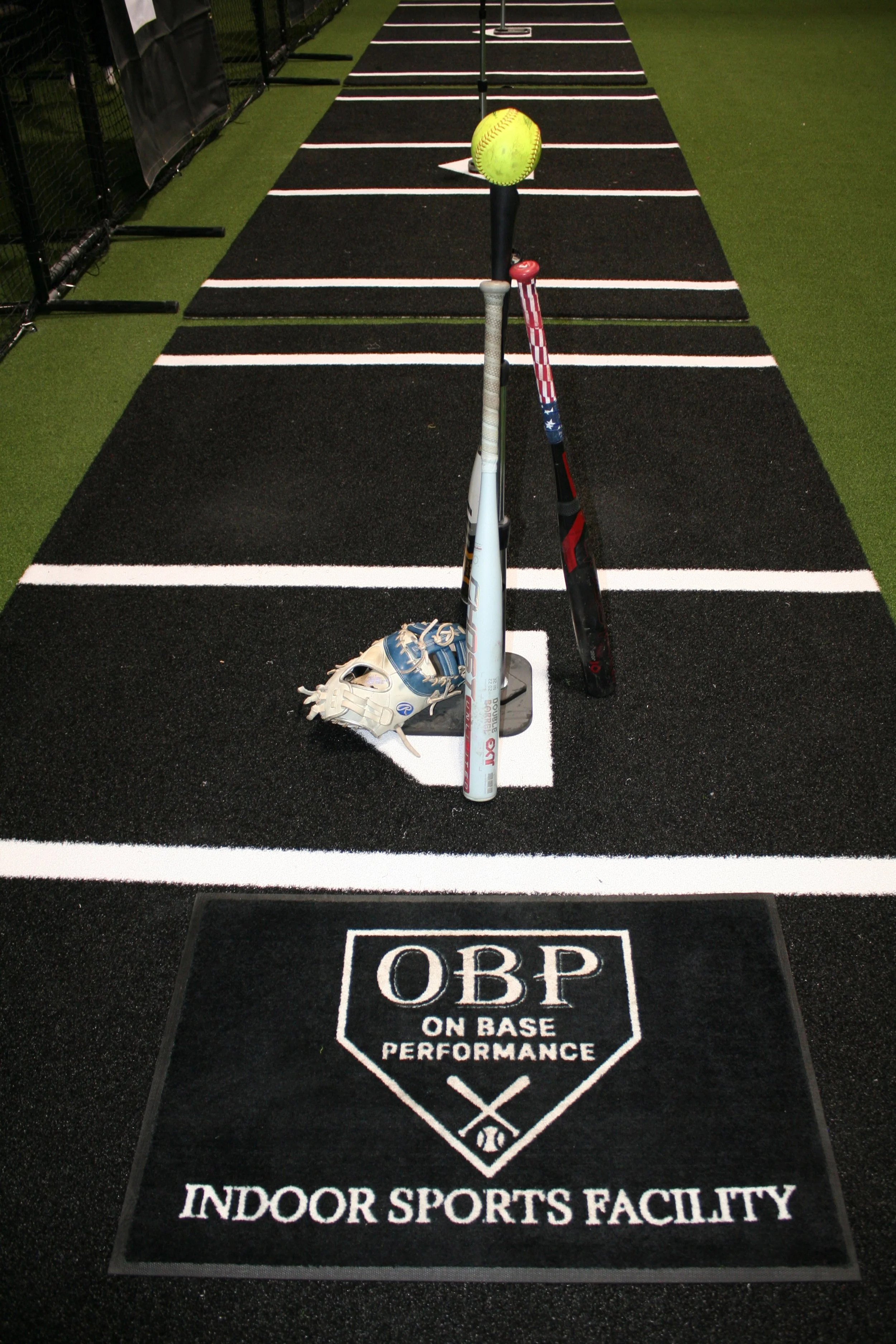 A baseball glove, two baseball bats, and a yellow softball on a black indoor sports facility mat with white lines and an OBP On Base Performance logo.
