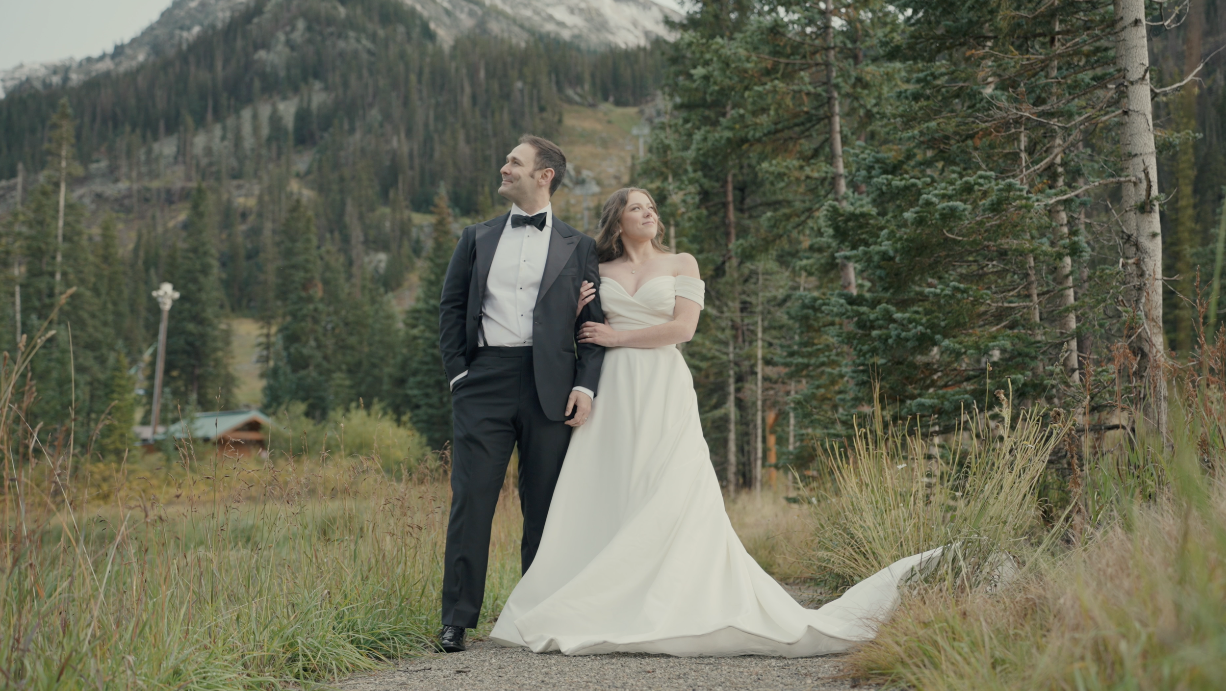 A newlywed couple standing outdoors in a forested mountain area during daytime, with the man in a tuxedo and the woman in a white wedding gown, looking happy.