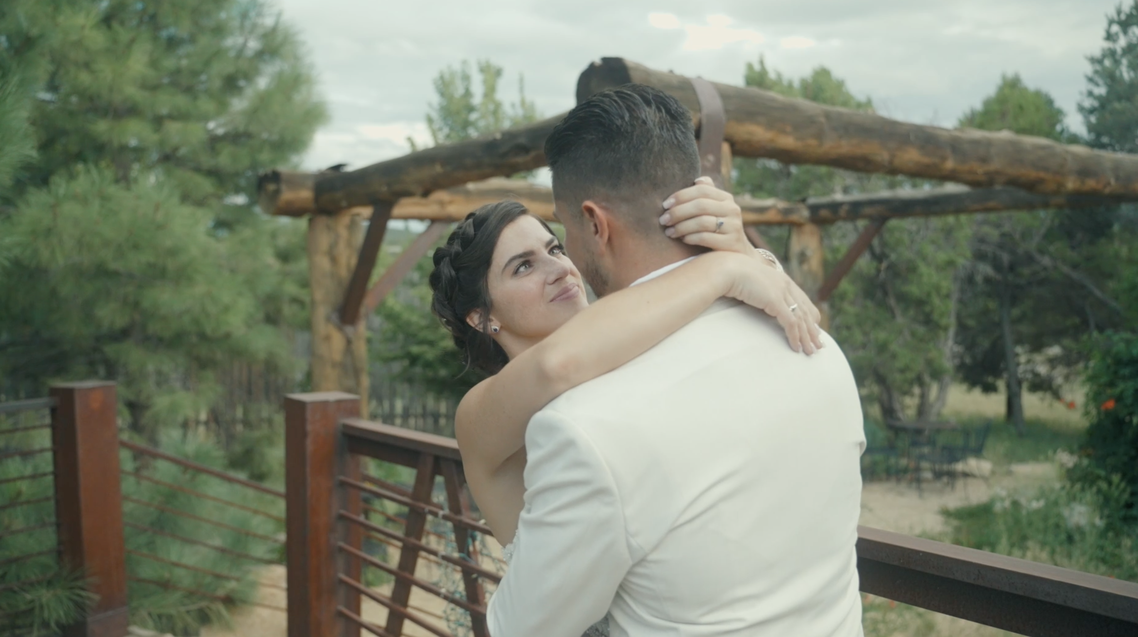 A bride and groom embrace outdoors on a wooden deck surrounded by green trees, with a cloudy sky overhead.
