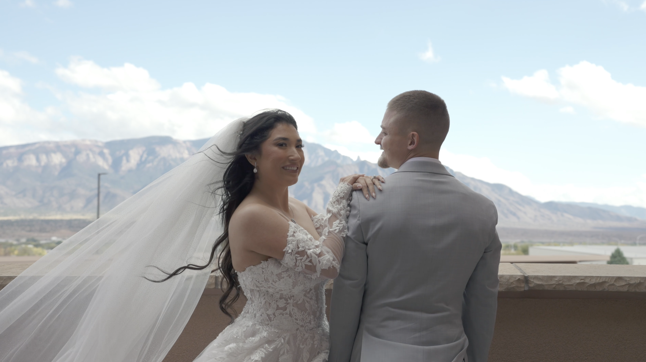 A bride and groom are sharing a dance outdoors with mountains in the background, the bride is wearing a lace wedding dress and veil, and the groom is in a gray suit.
