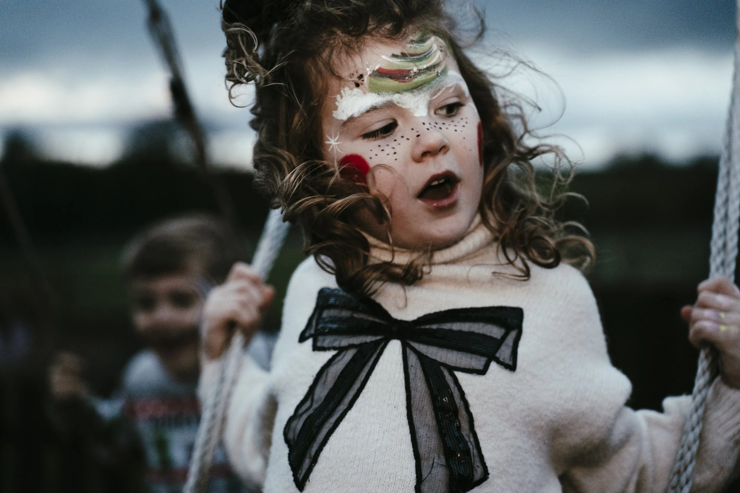 A young girl with face paint resembling a rainbow, stars, and dots, wearing a cream sweater with a black bow, riding a swing outdoors during dusk, with a boy in the background.