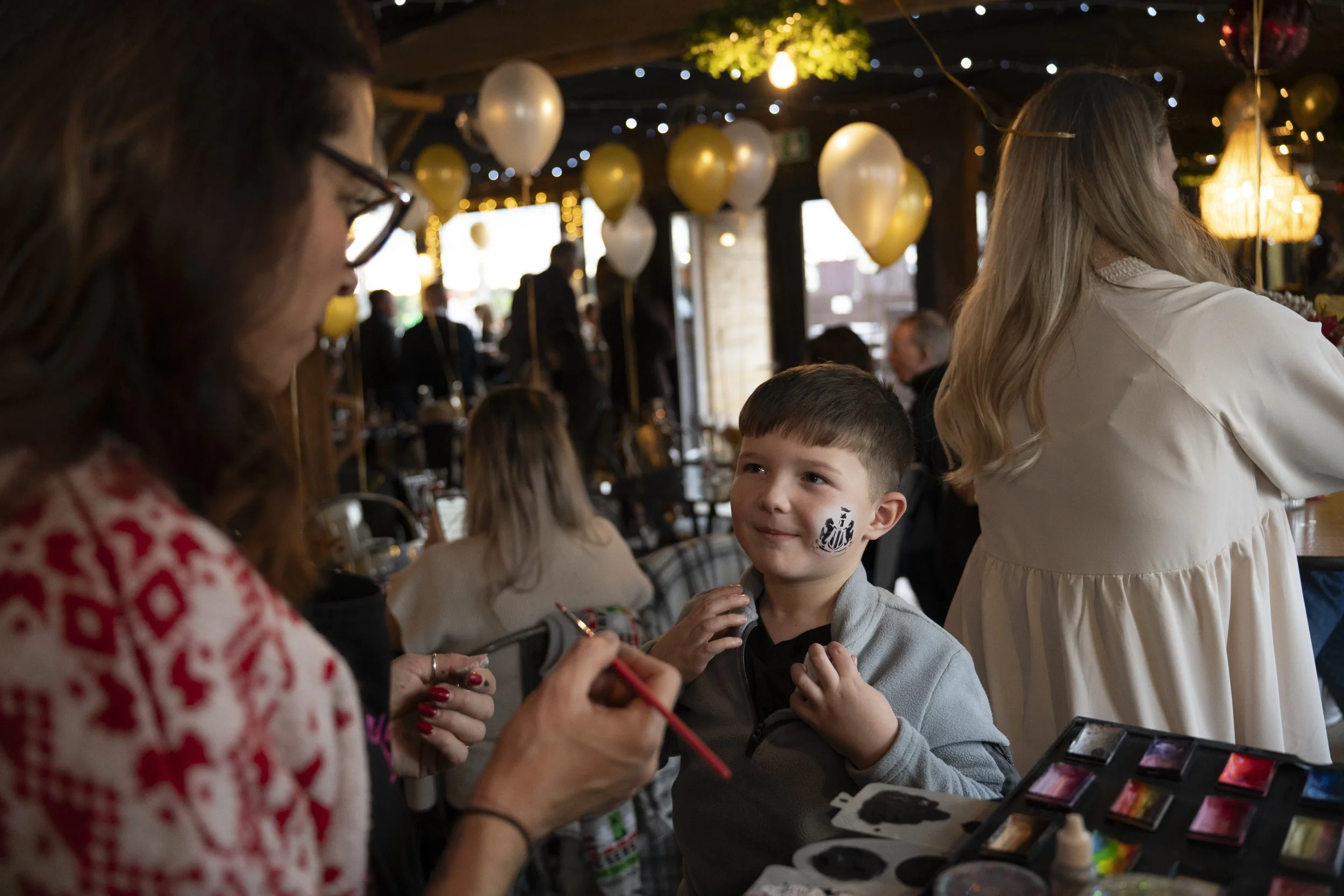 A young boy having his face painted by a woman at a festive indoor event decorated with balloons and string lights.