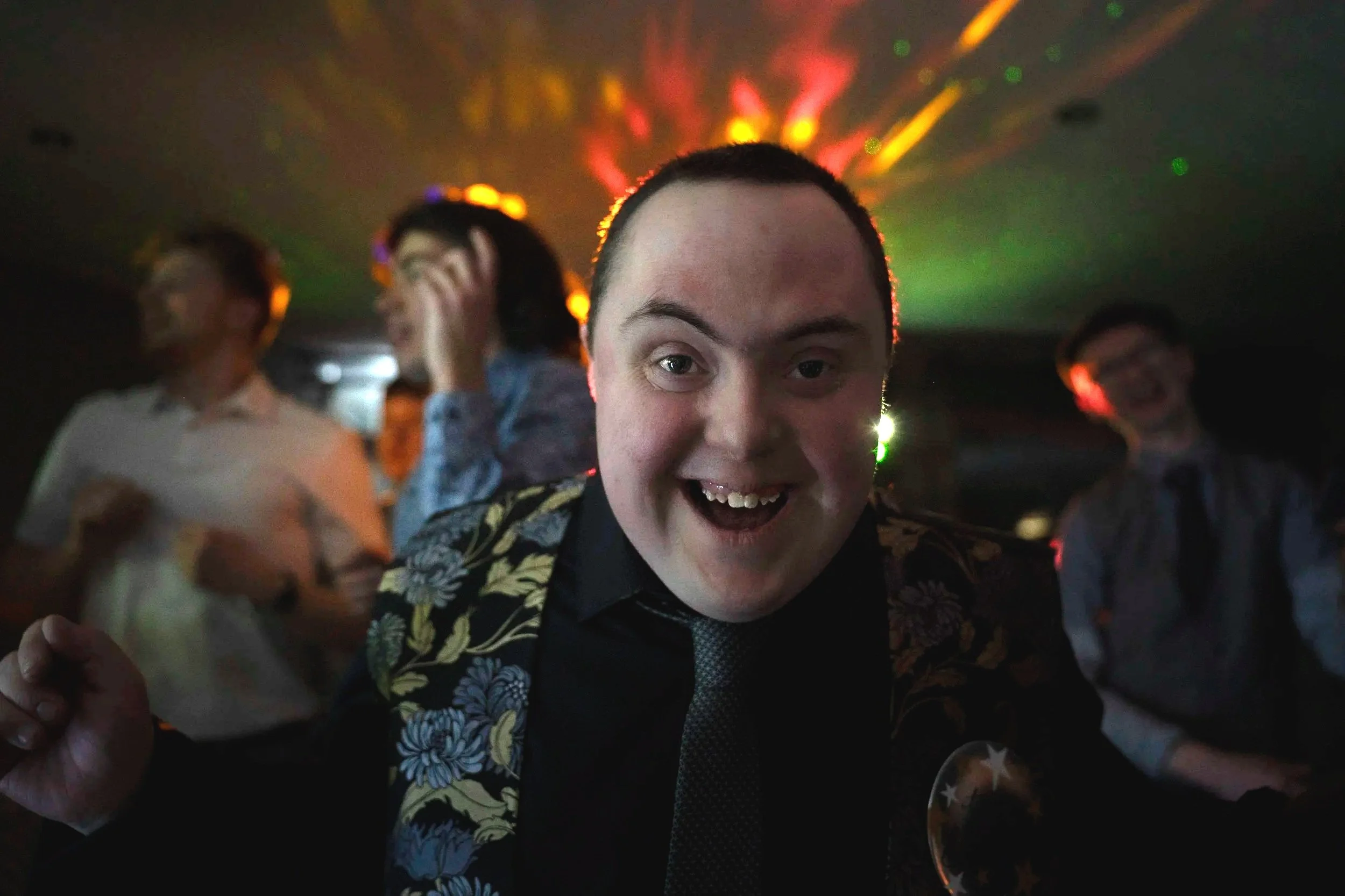 A man smiling and celebrating at a party or dance event, with colorful disco lights illuminating the background and other people dancing or socializing.