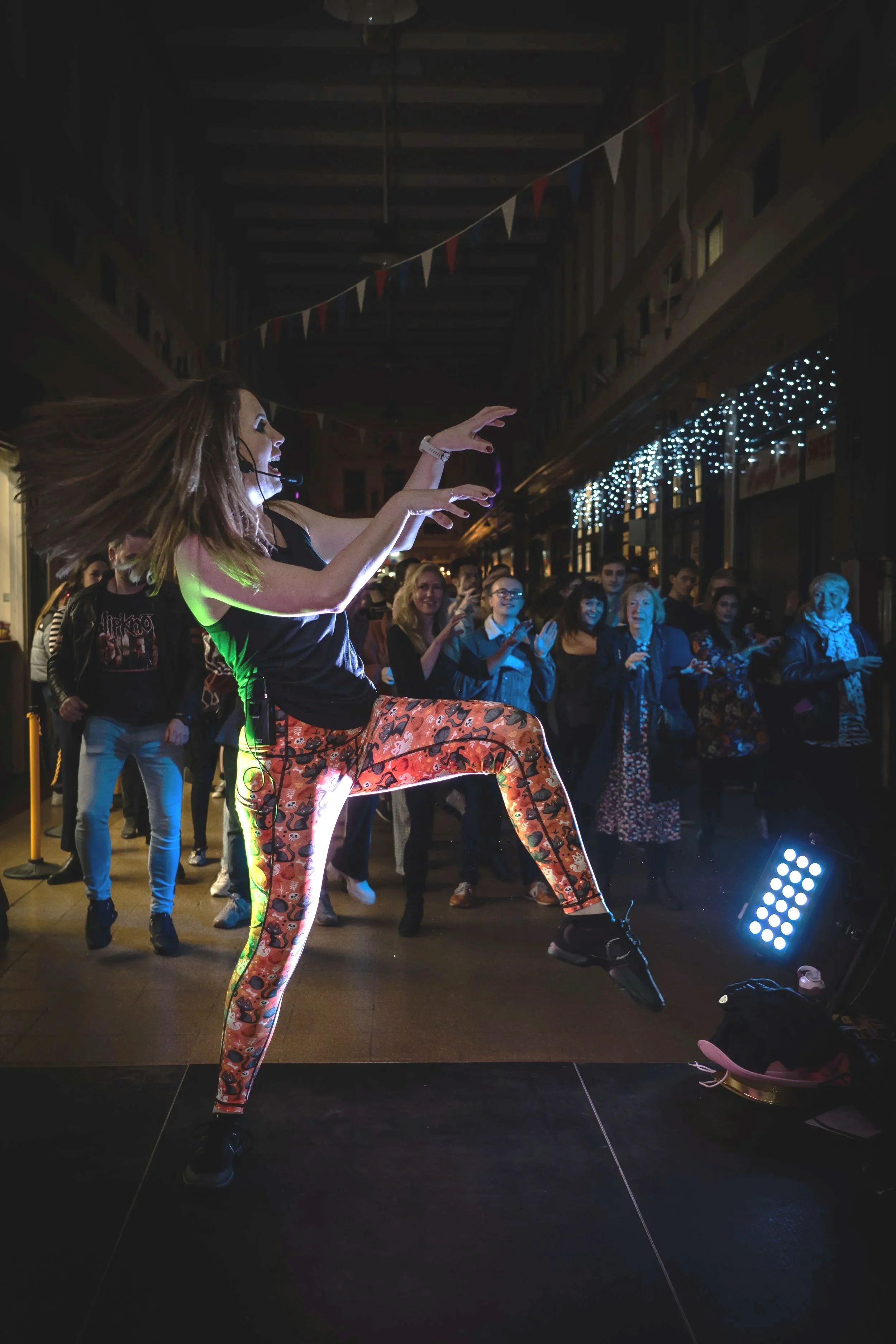 A woman in colorful leggings and a black top performing as a dancer on stage during a lively dance event with audience members watching and cheering in a dimly lit venue with festive string lights.