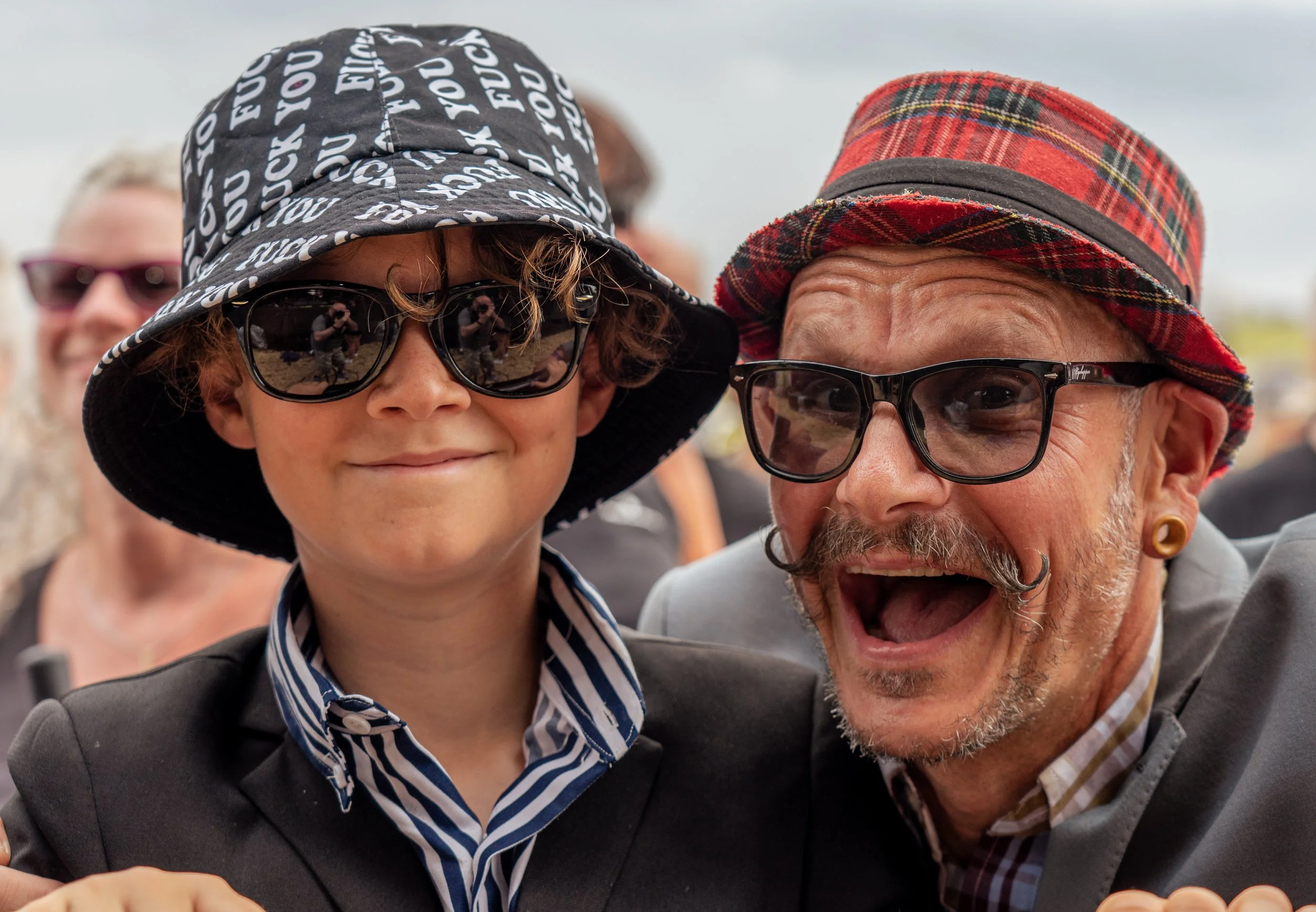 A woman and a man wearing sunglasses and stylish hats, smiling and taking a selfie together at an outdoor event.