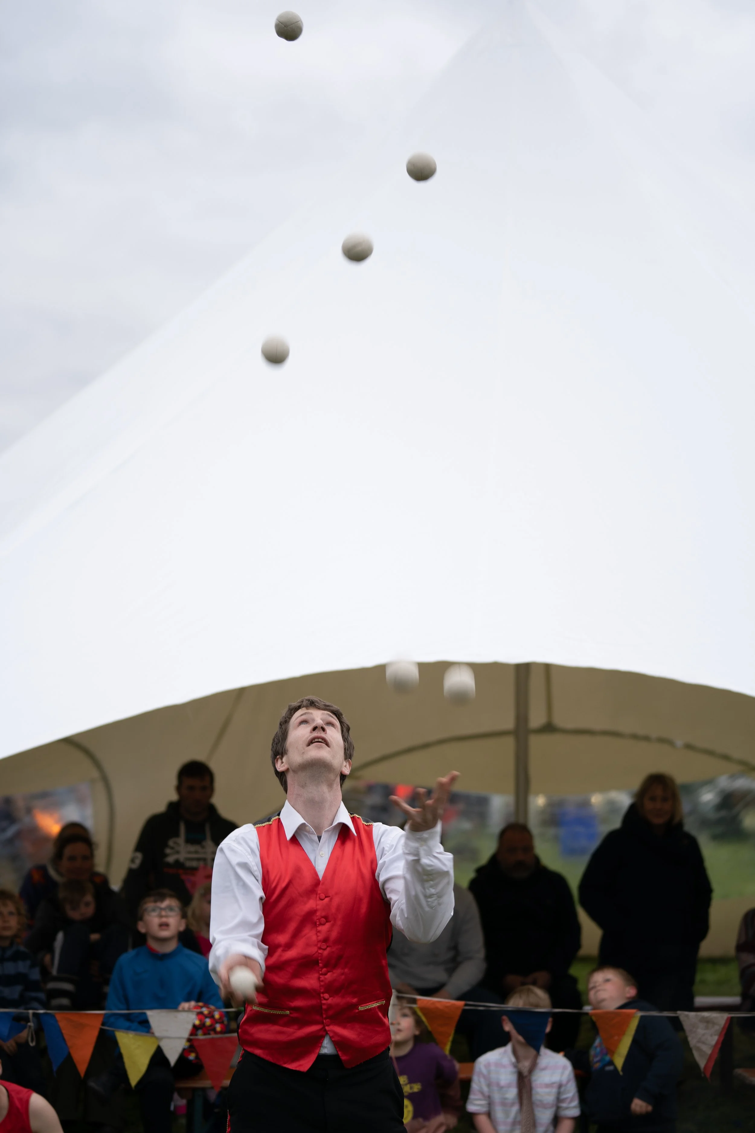 A clown juggles balls in front of a crowd under a tent at an outdoor event.