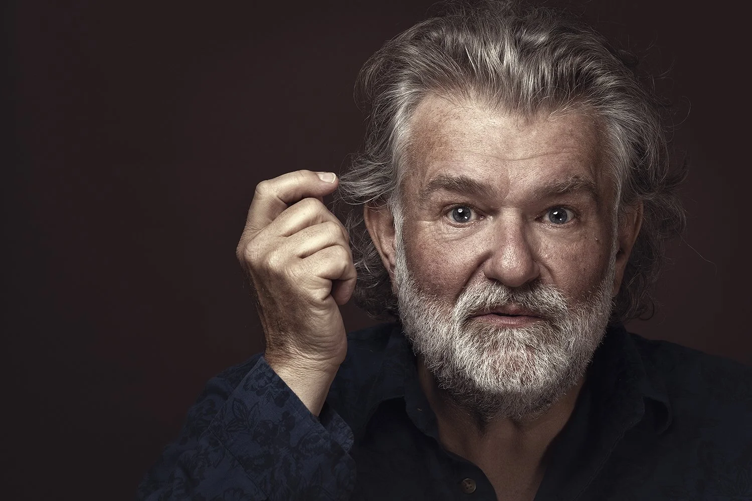 A middle-aged man with gray hair and a beard, holding a lock of his hair, looking directly at the camera against a dark background.