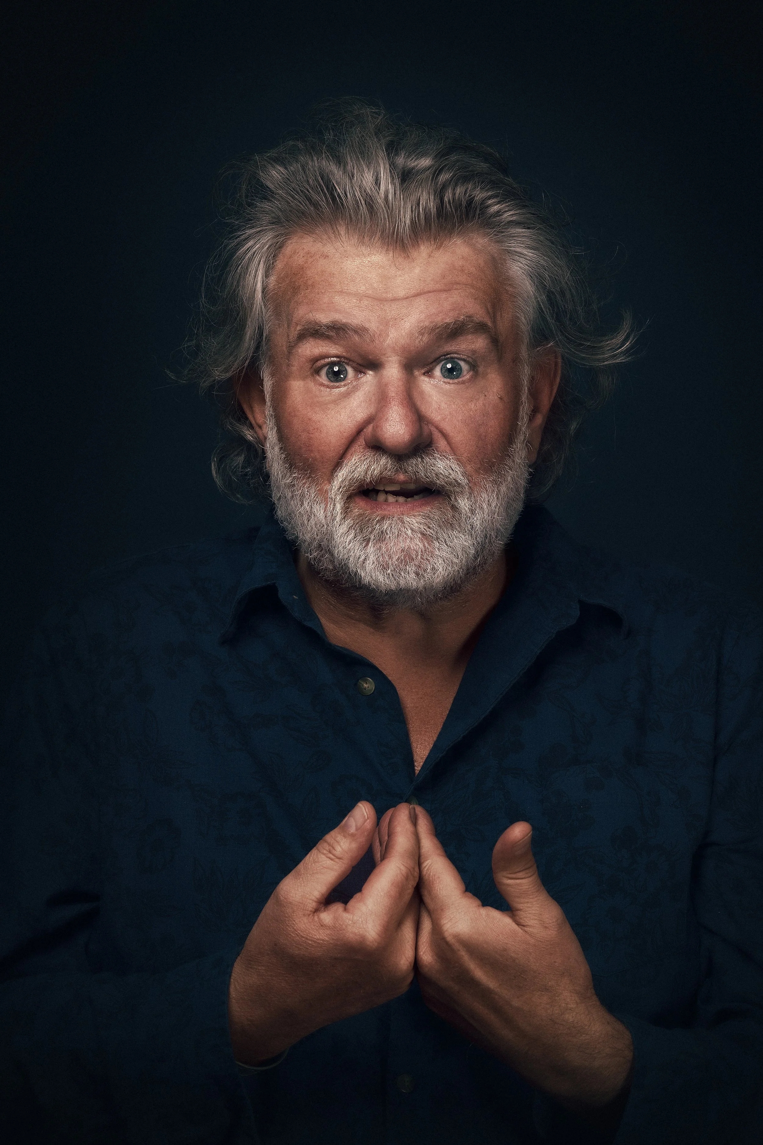 An older man with gray hair and a beard, wearing a dark blue patterned shirt, looking surprised and holding the top of his shirt with both hands against a dark background.