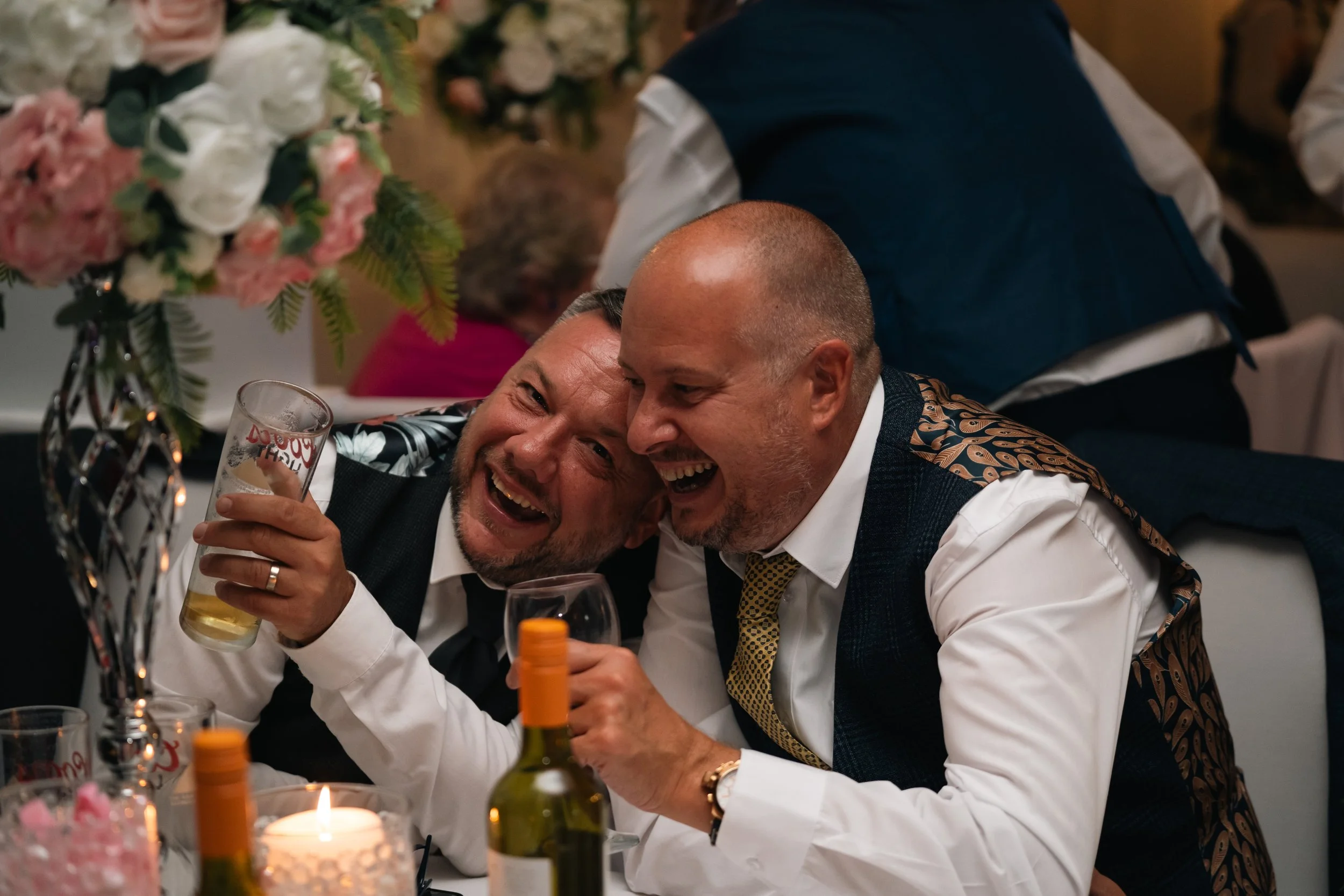 Two men laughing and celebrating at a dinner party, holding drinks, with flower arrangements and a lit candle on the table.