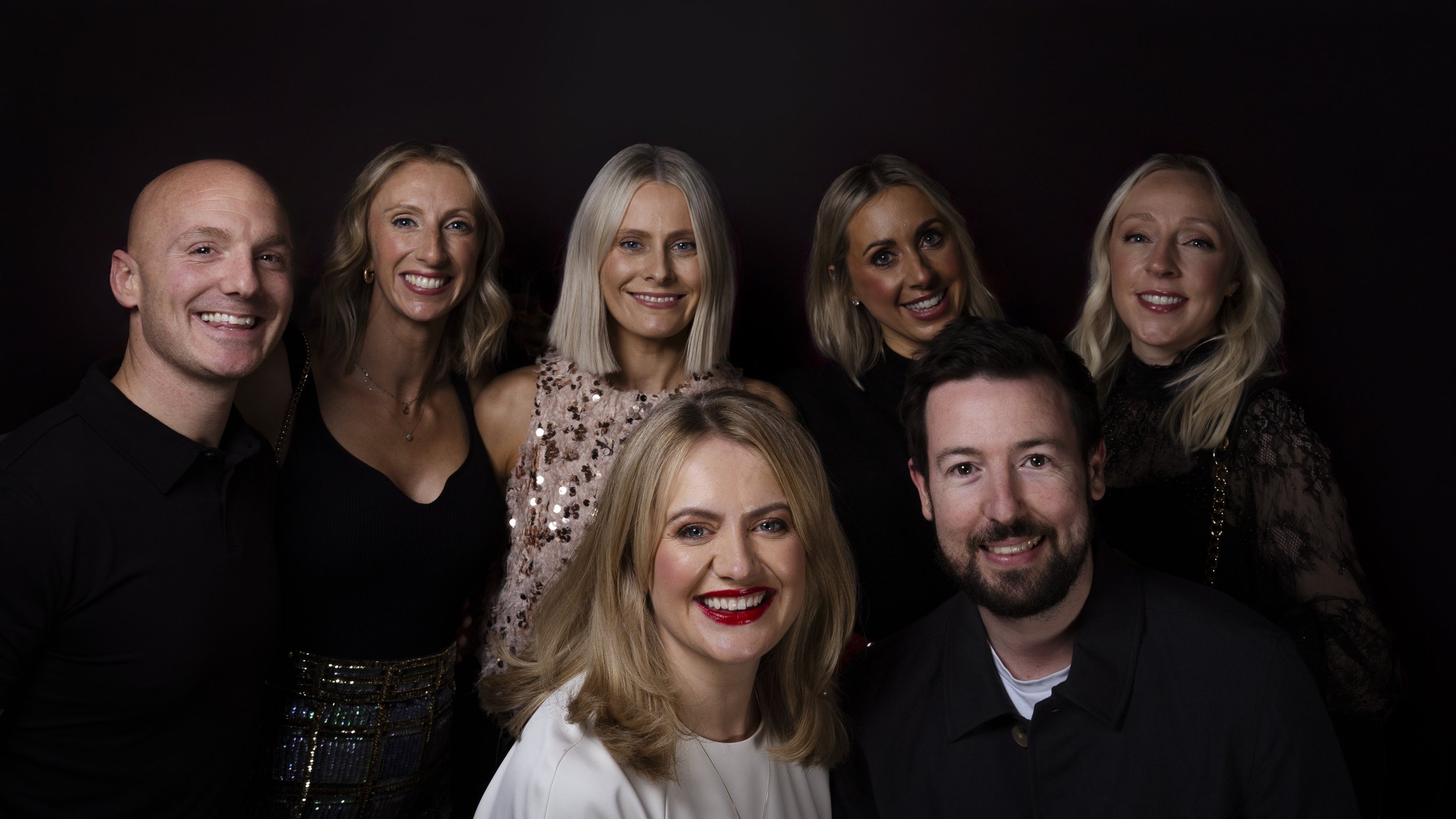 Group of seven diverse adults posing for a photo against a dark background, smiling, dressed in semi-formal attire.