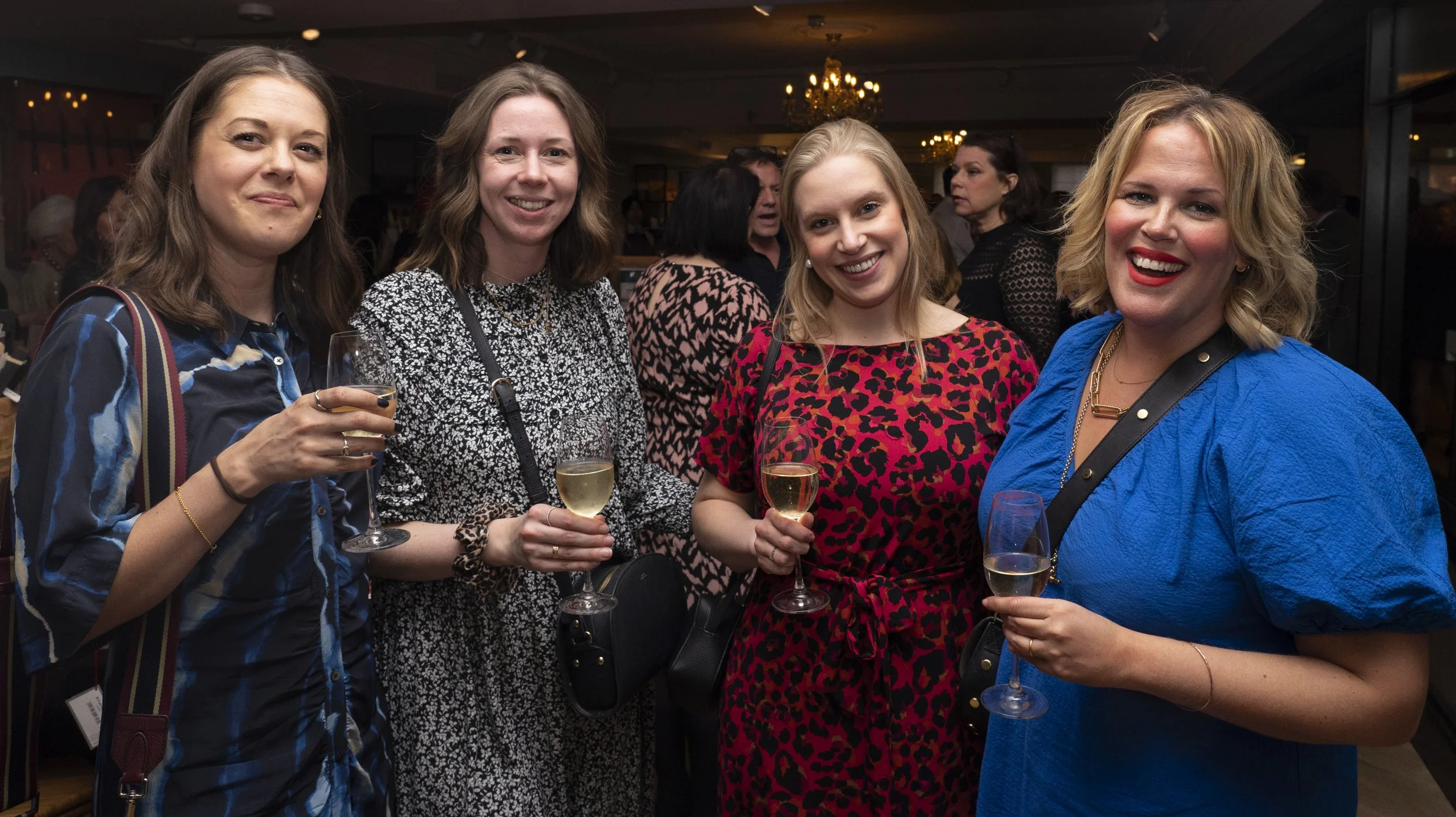 Four women smiling and holding glasses of white wine at an indoor social event.