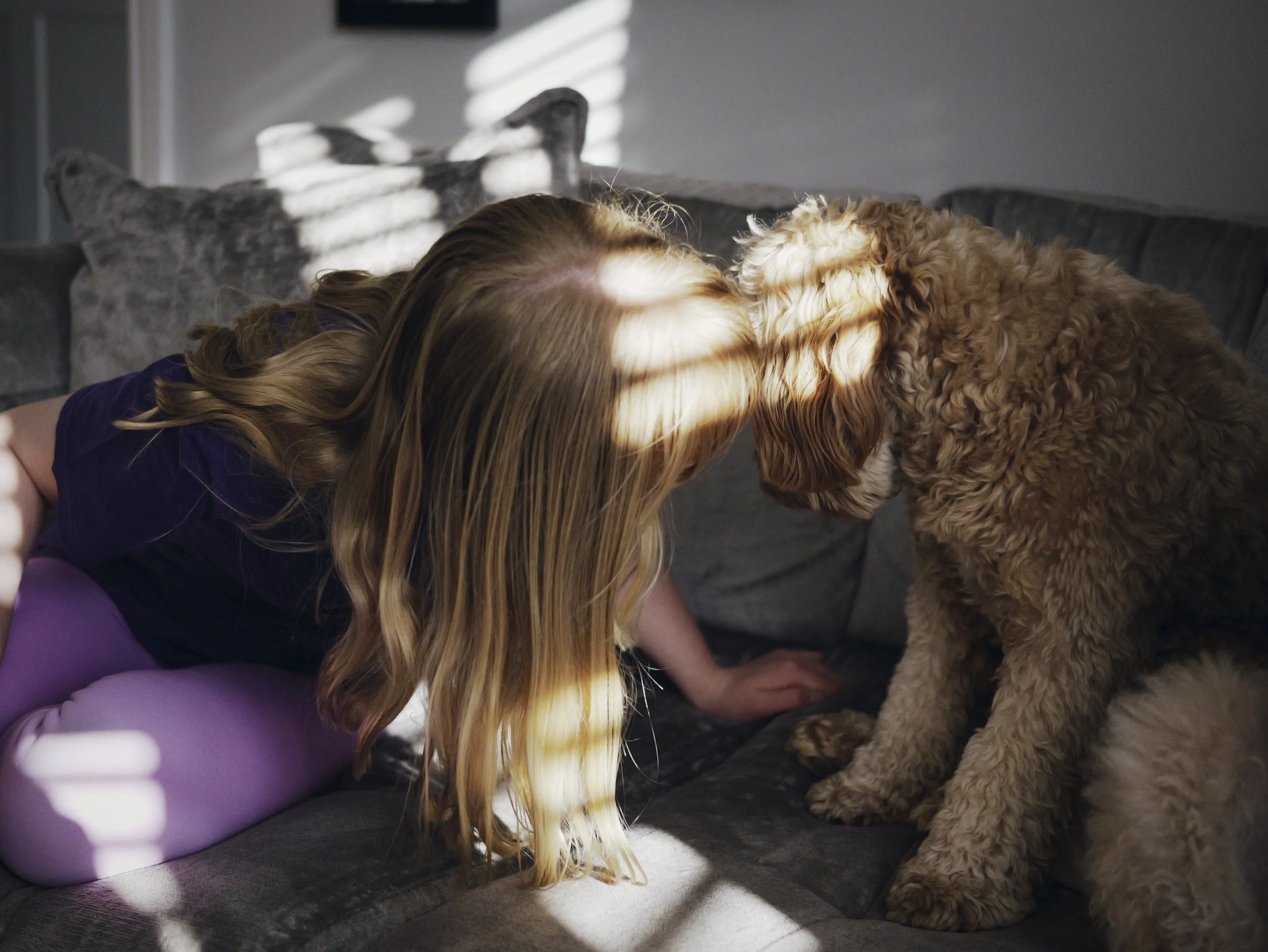 A girl with long, wavy blonde hair leaning forward to kiss a large, fluffy, light brown dog on the nose, sitting on a dark sofa in a living room.