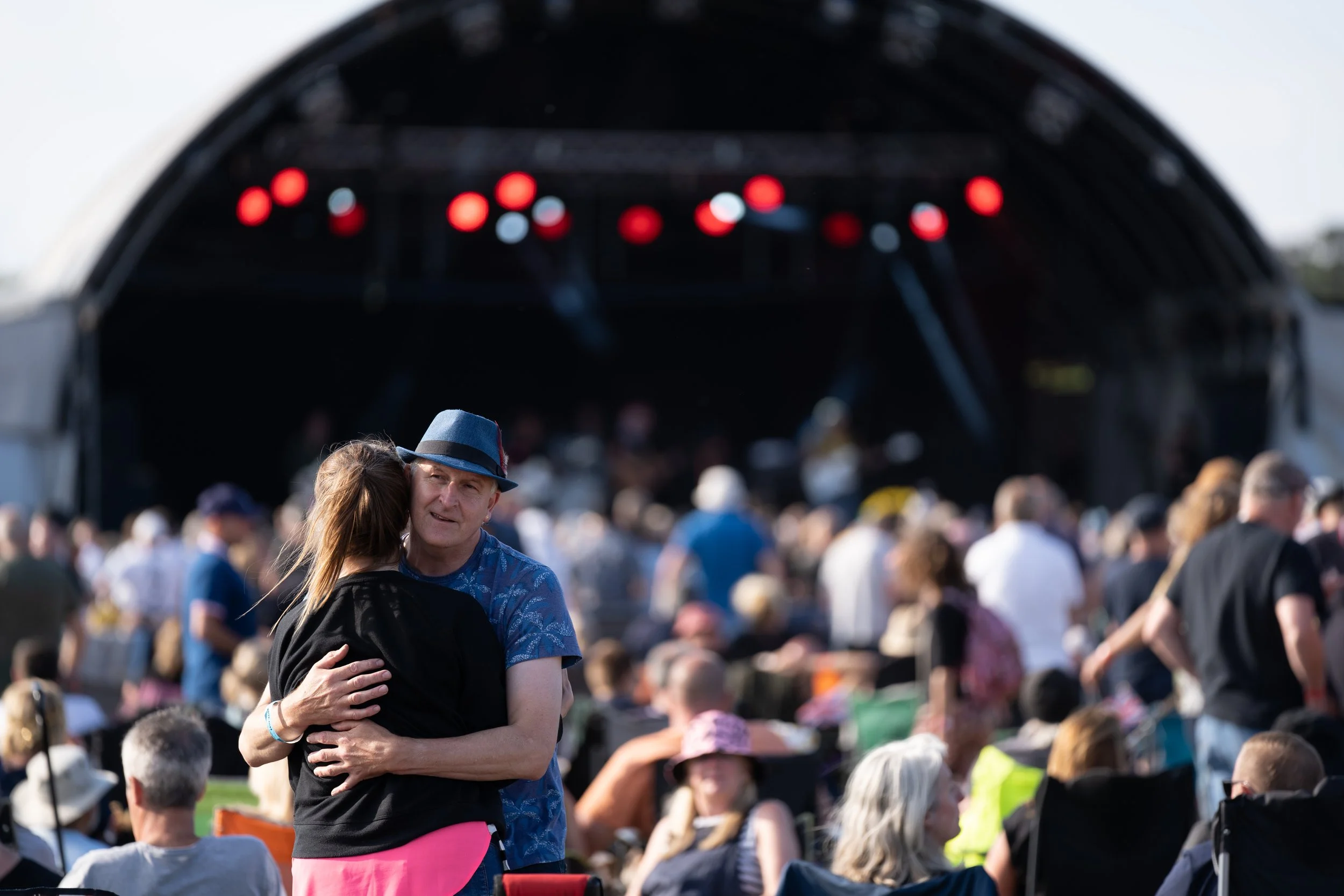 A man and woman hugging at an outdoor concert or festival with a stage and crowd in the background.