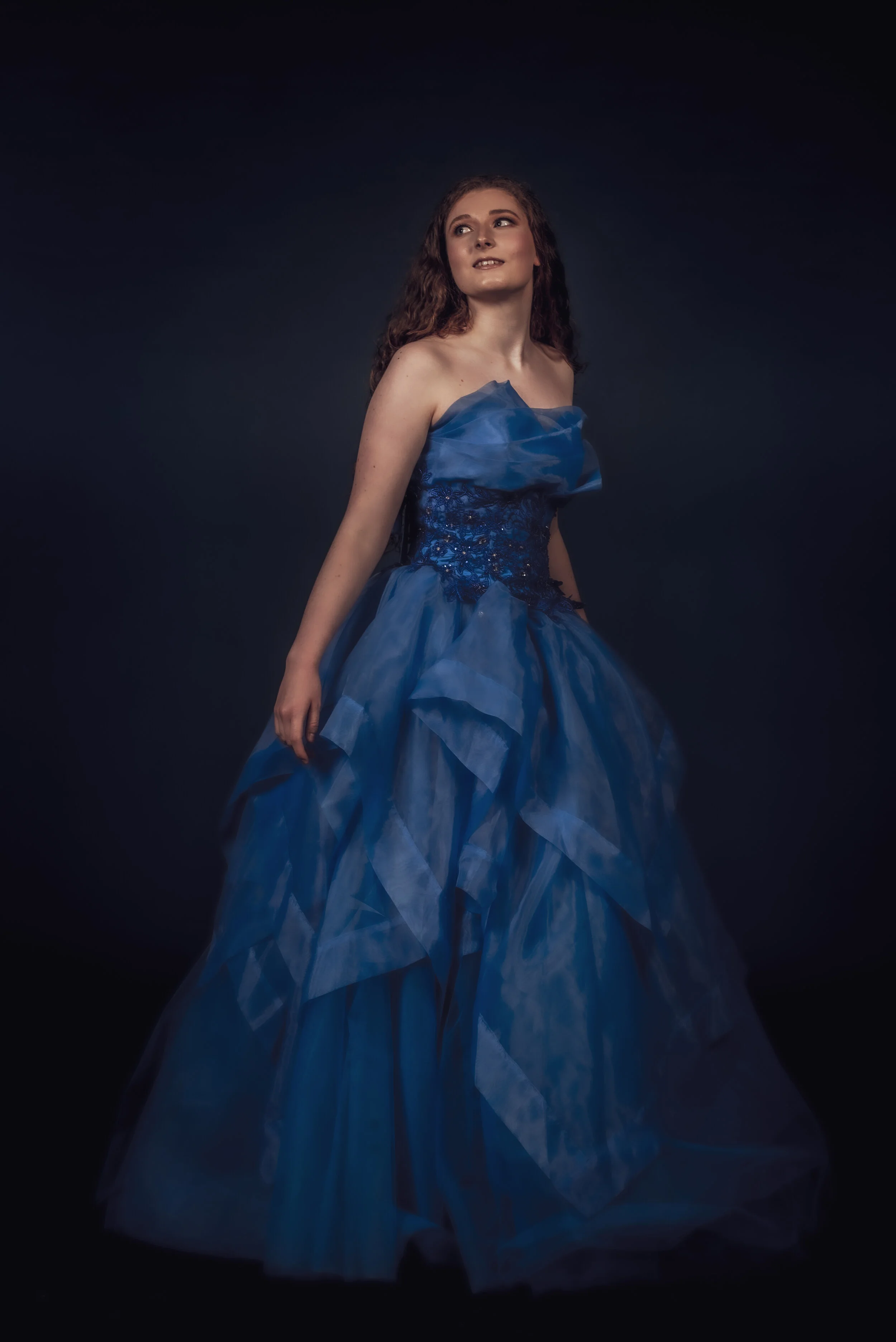 A young woman in an elegant, strapless blue gown with ruffled layers standing against a dark background.