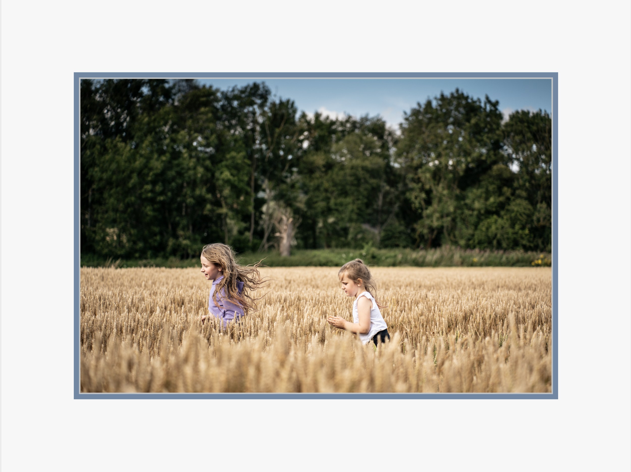 Two young girls playing in a wheat field with trees in the background on a sunny day.
