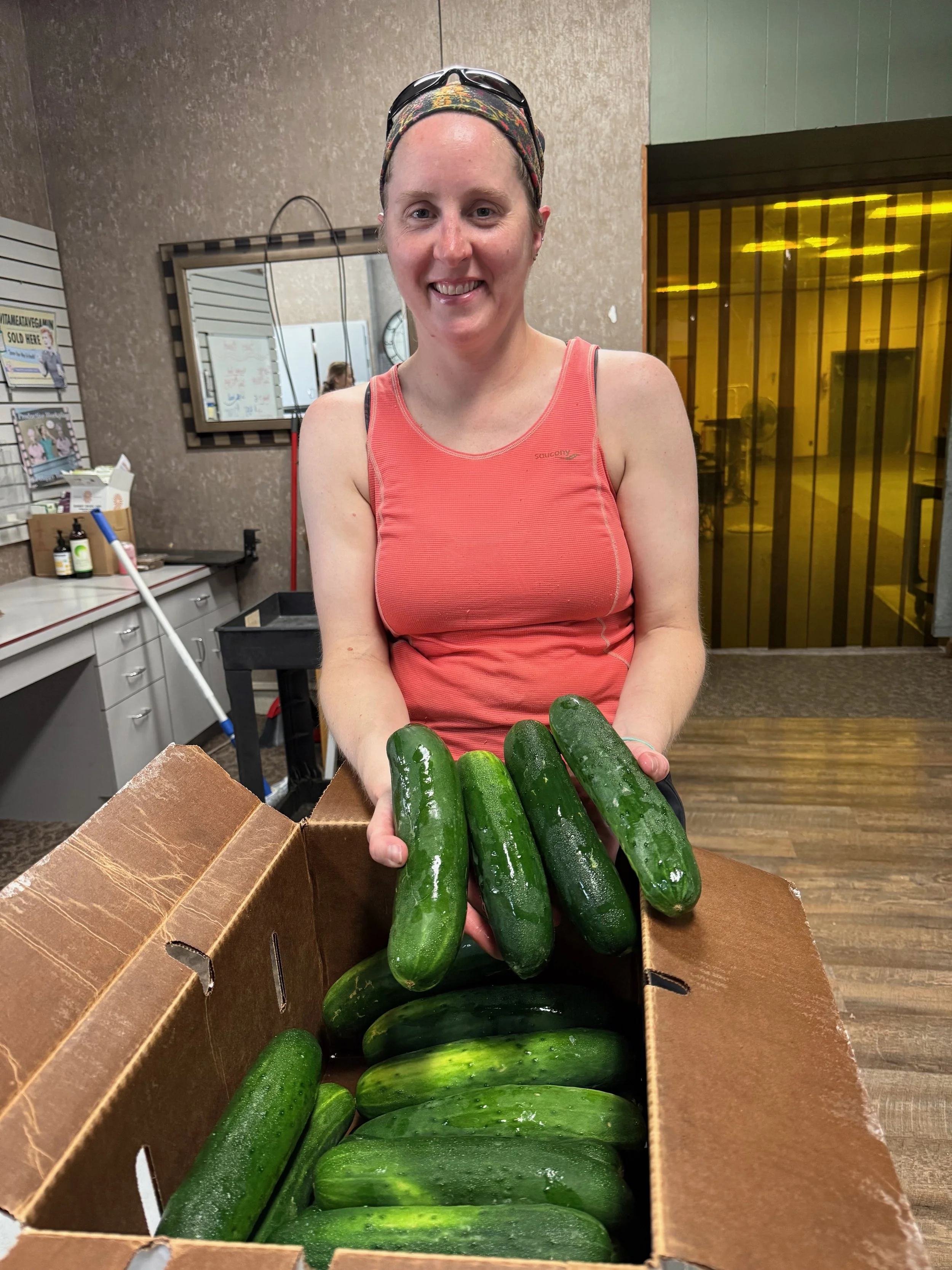 A woman with a headband and sunglasses on her head holding fresh cucumbers inside a cardboard box.
