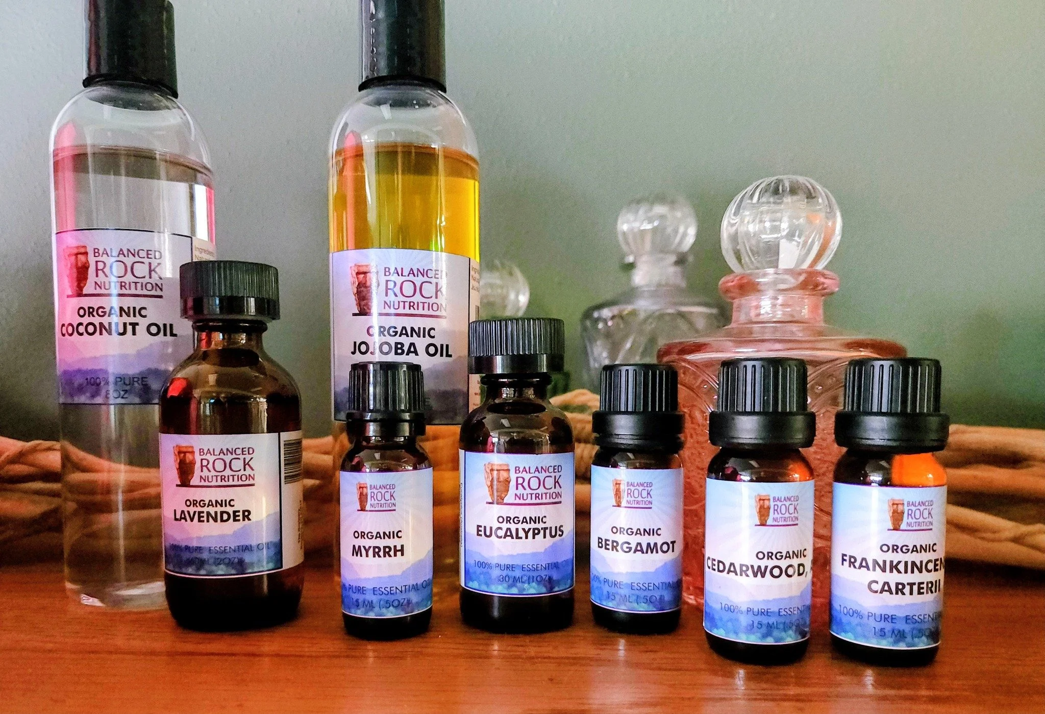 A collection of essential oils in various small bottles, including lavender, myrrh, eucalyptus, bergamot, cedarwood, frankincense, and caper, along with larger bottles labeled coconut oil, jojoba oil, and another with a yellow liquid, arranged on a wooden surface.