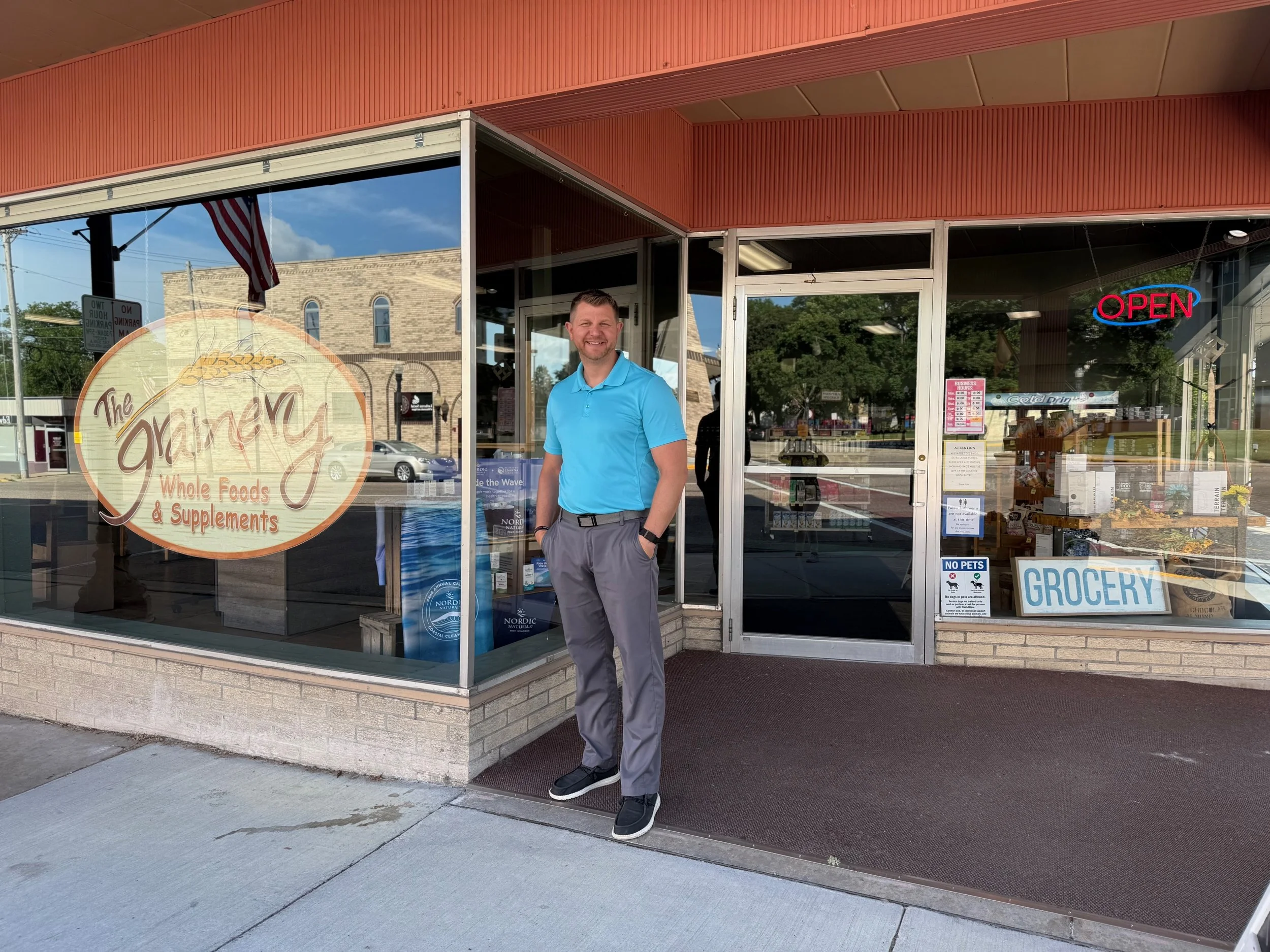 A man in a blue polo shirt and gray pants standing outside a store with a sign that reads "The Grain" Whole Foods & Supplements. The store has large glass windows, an 'Open' sign, and various notices on the door. Reflection of a building, trees, and parked cars is visible in the windows.