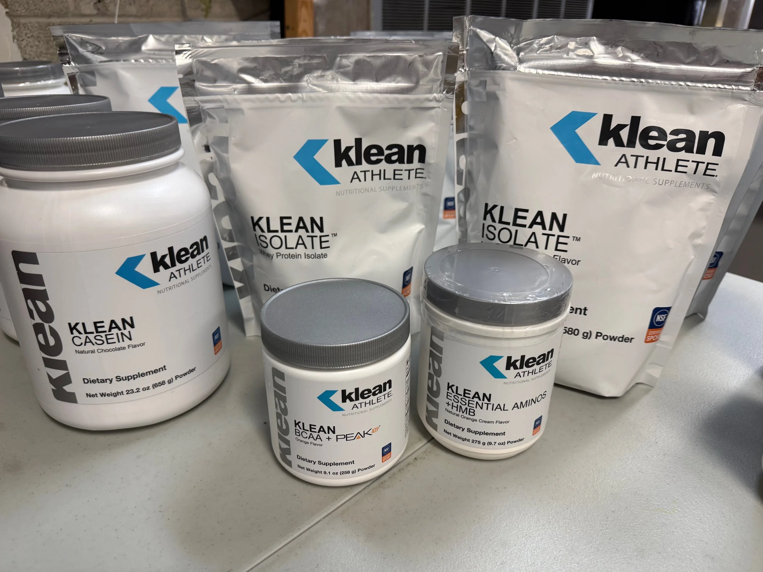 Assorted nutritional supplements in white containers and silver pouches with blue and black labels, including whey protein isolate, casein, essential amino acids, BCAA, orange flavor, and other health supplements.