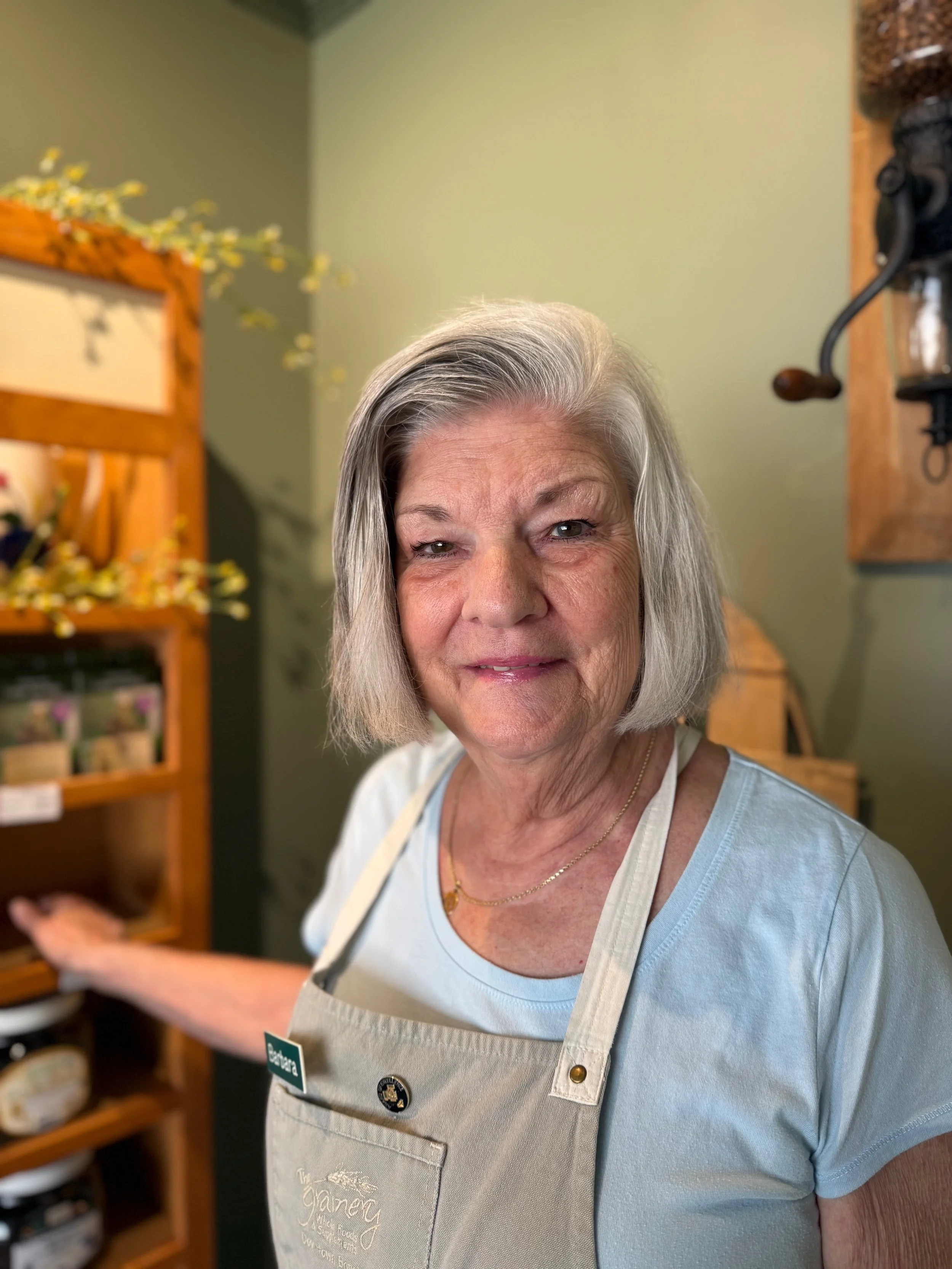 A smiling elderly woman wearing a light blue t-shirt and a beige apron with a name tag, standing in a cozy kitchen with wooden shelves filled with jars and decorative items.