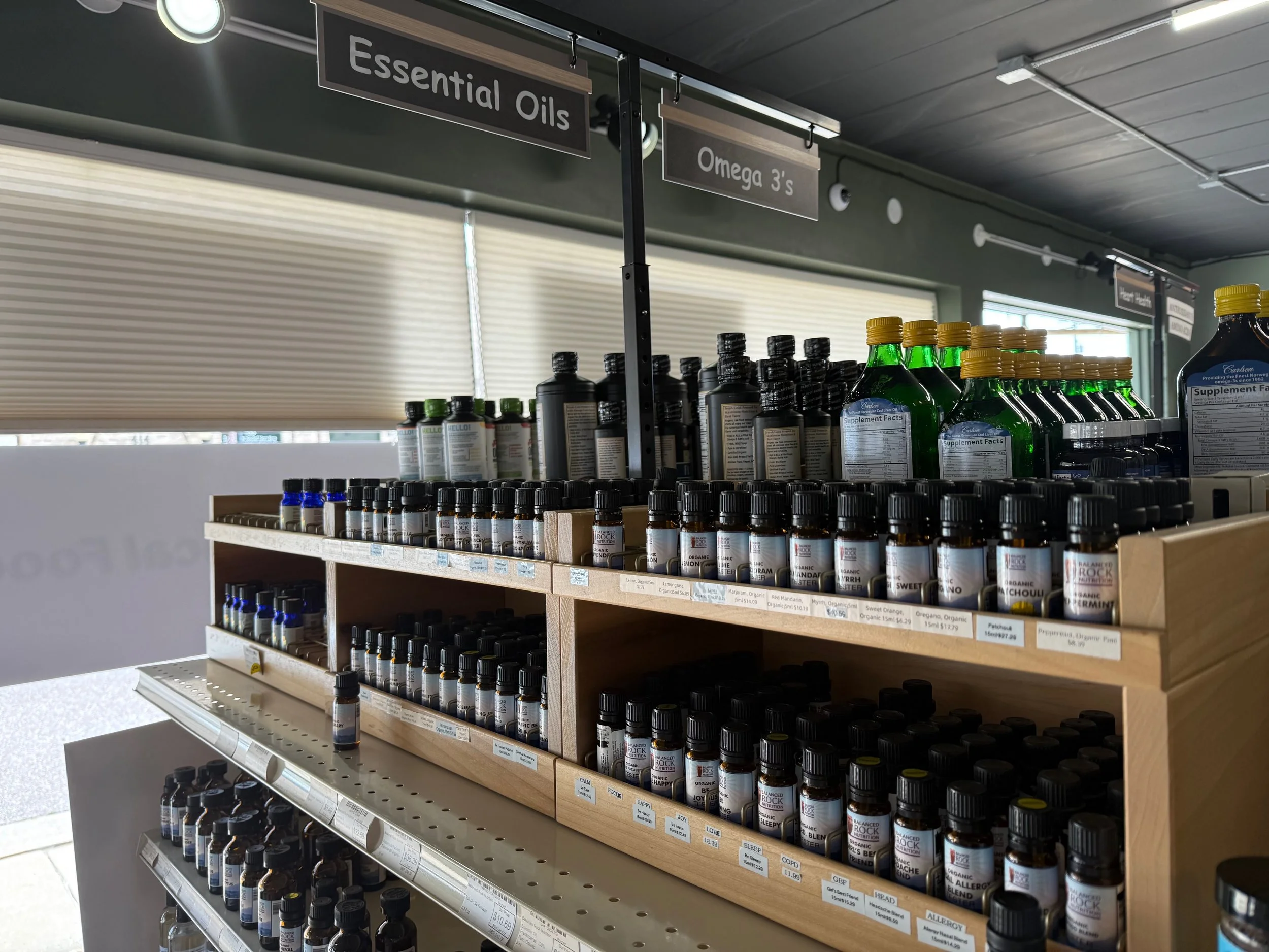 A retail display shelf holding essential oils and omega 3 supplements. The wooden shelves are filled with small bottles, organized with labels indicating product types and prices. Signage above reads 'Essential Oils' and 'Omega 3's'.