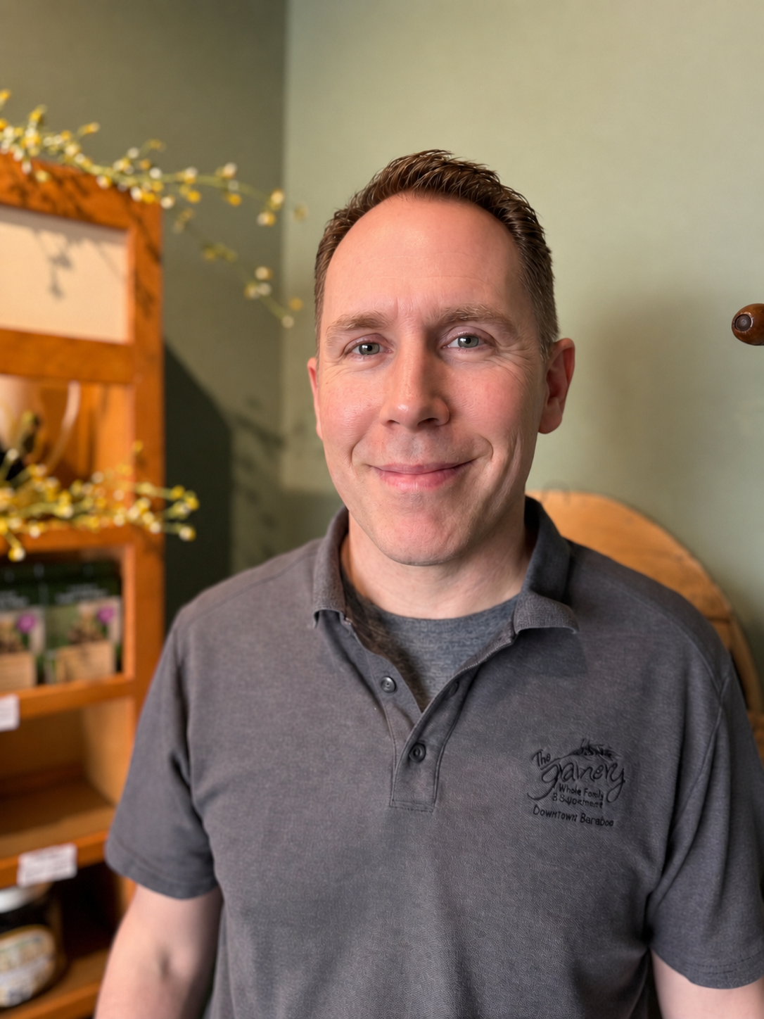 A man with short brown hair and blue eyes smiling in front of a green wall, wearing a gray polo shirt with a logo that says "The Gallery". There are wooden shelves with flowers and potted plants in the background.
