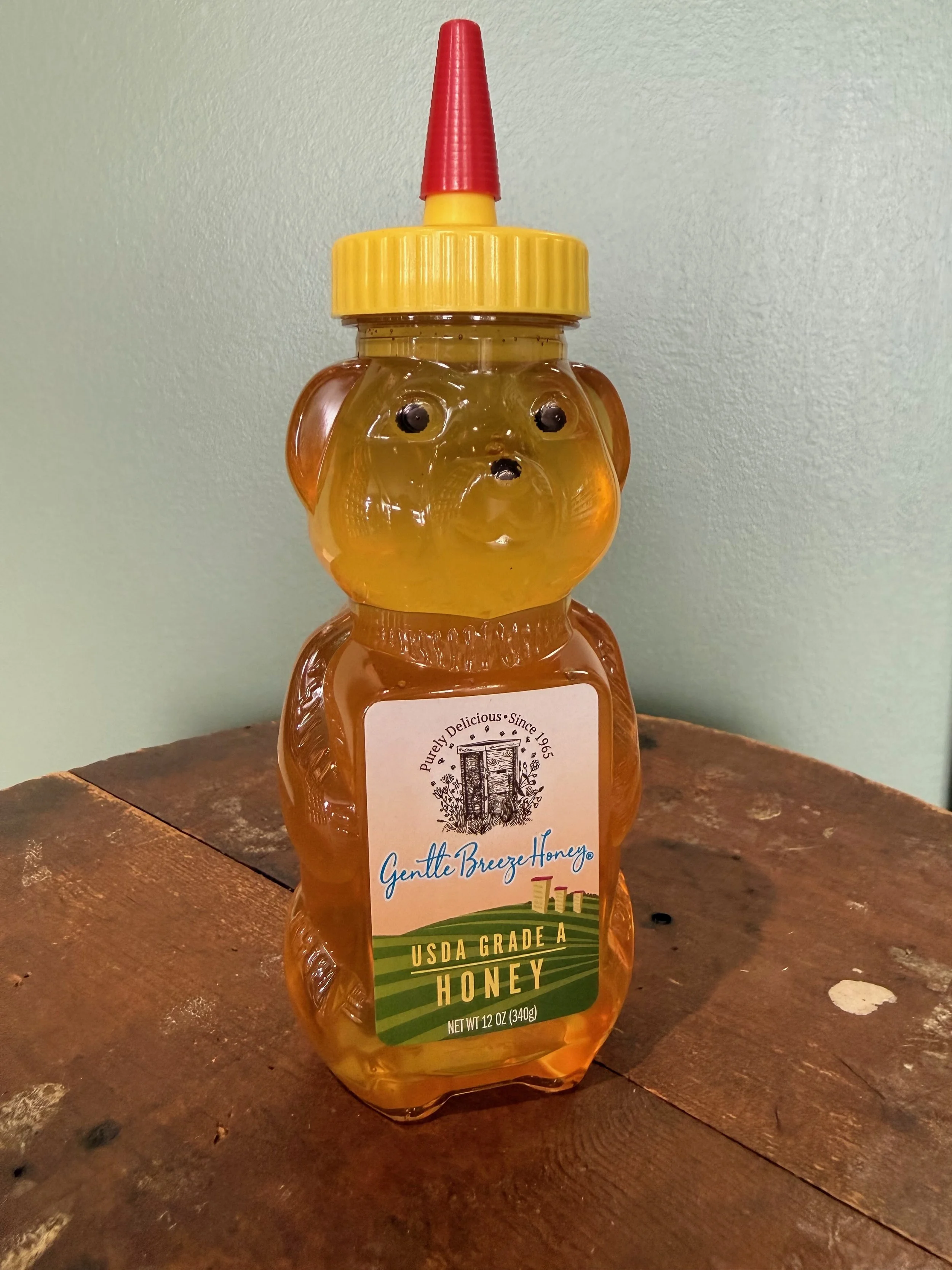 Bottle of honey shaped like a bear with a red squeeze top and yellow cap, labeled "Gentle Breeze Honey," sitting on a wooden surface.