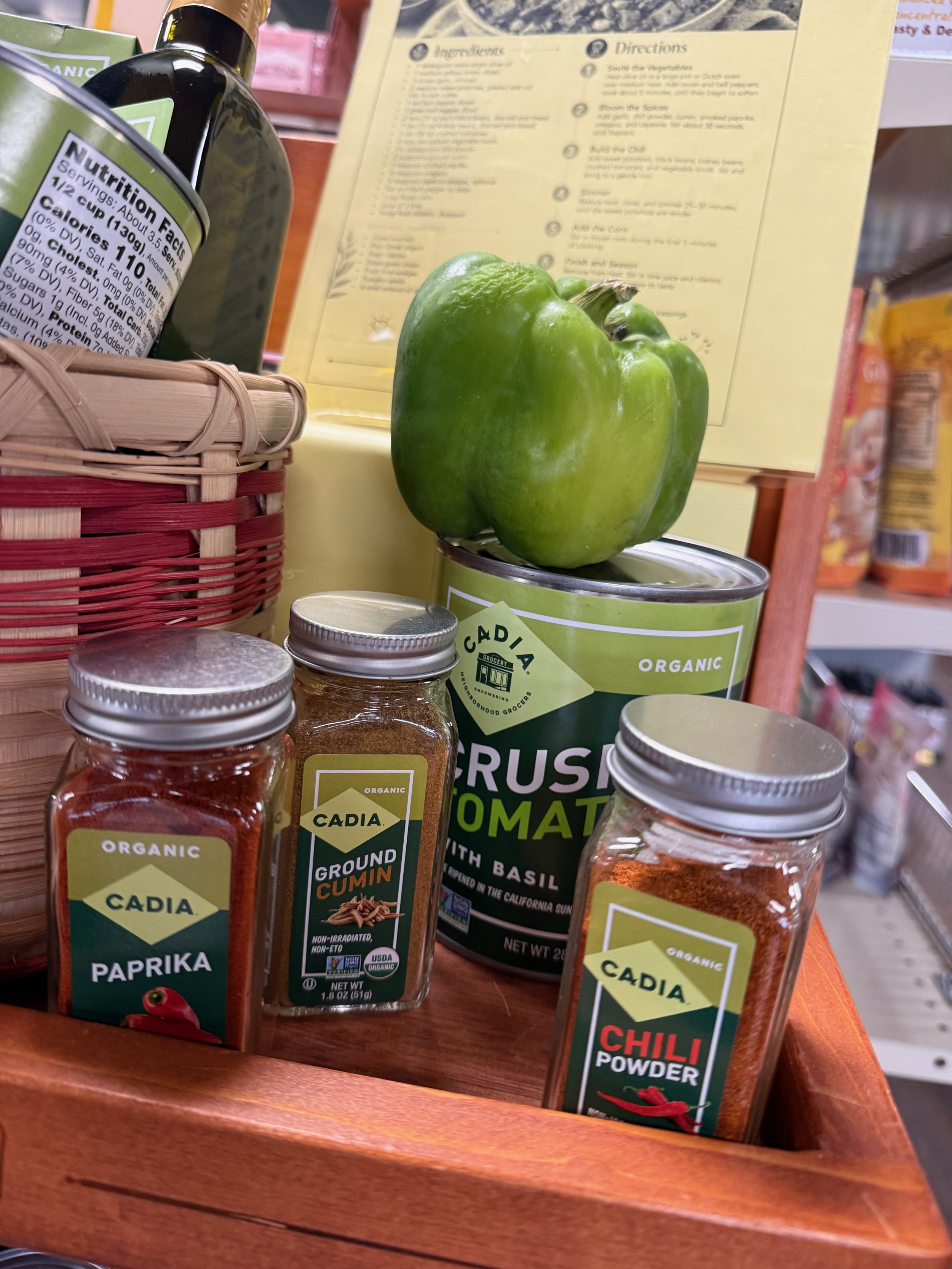 A green bell pepper on top of a can of crushed tomato, with jars of paprika, cumin, and chili powder surrounding them, in a wooden display.