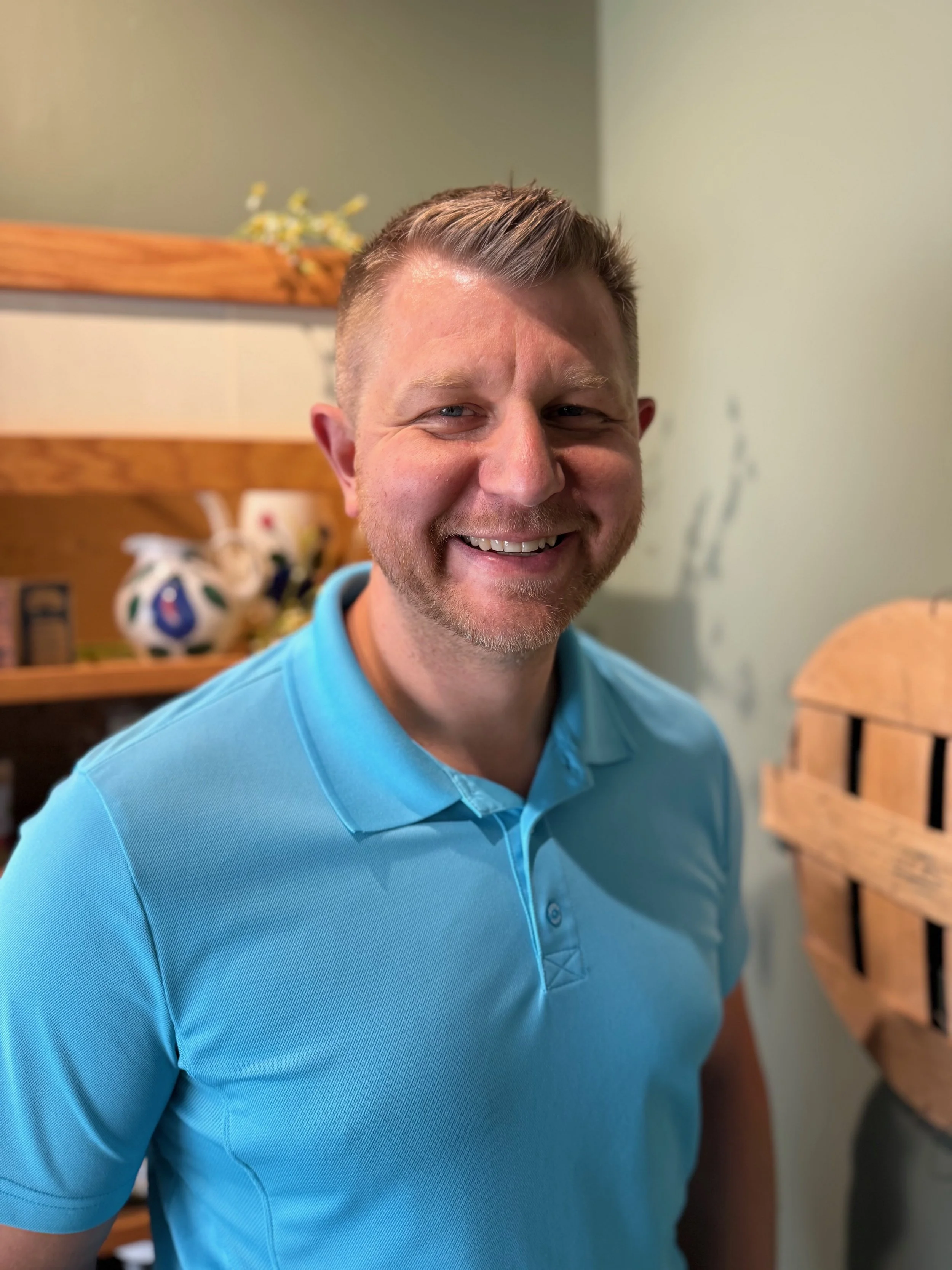 A smiling man with short blonde hair and wearing a light blue polo shirt, standing indoors in front of a wooden shelf with decorative items, against a light-colored wall.