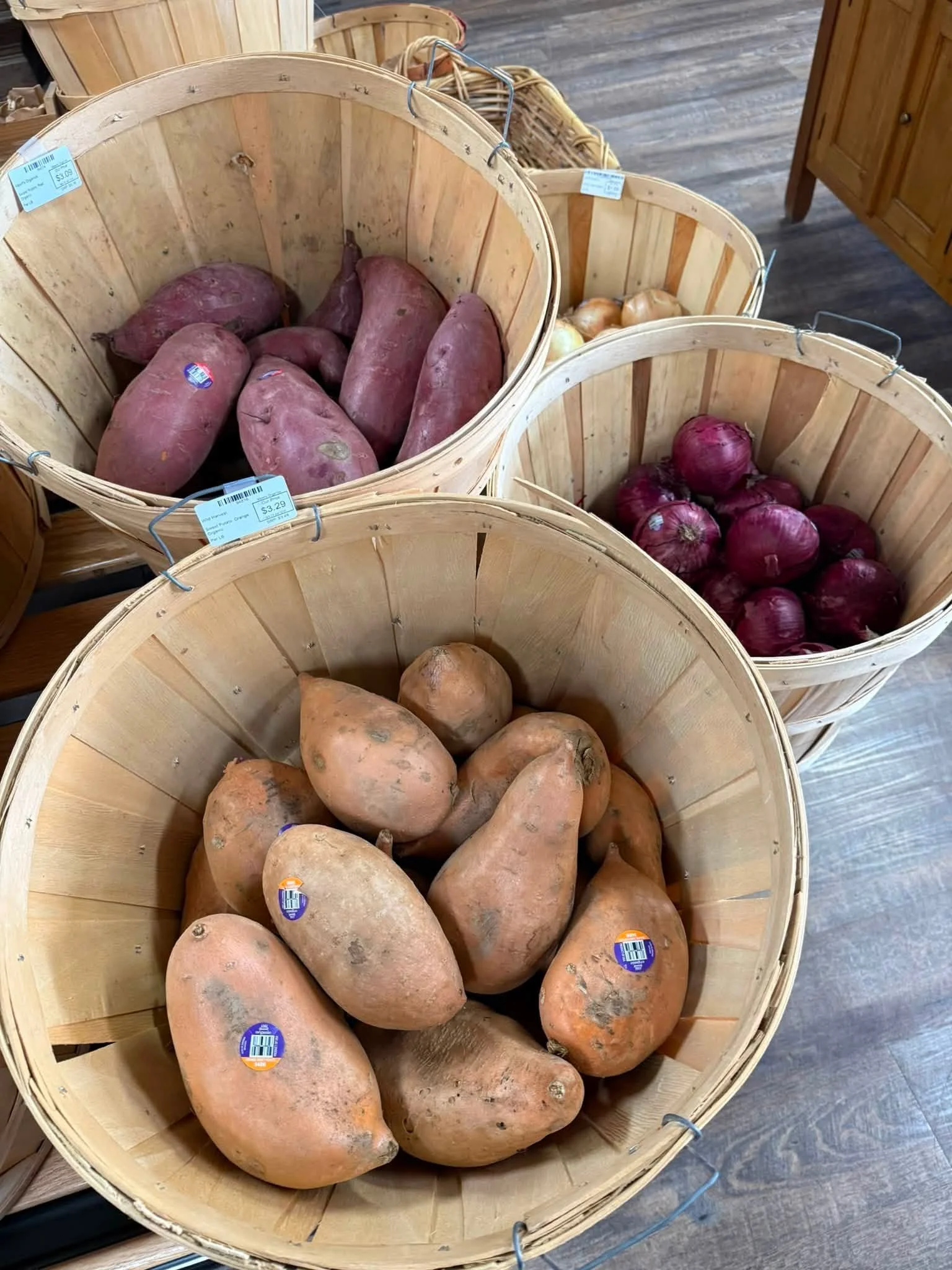 Multiple sweet potatoes and onions in wooden baskets at a grocery store.
