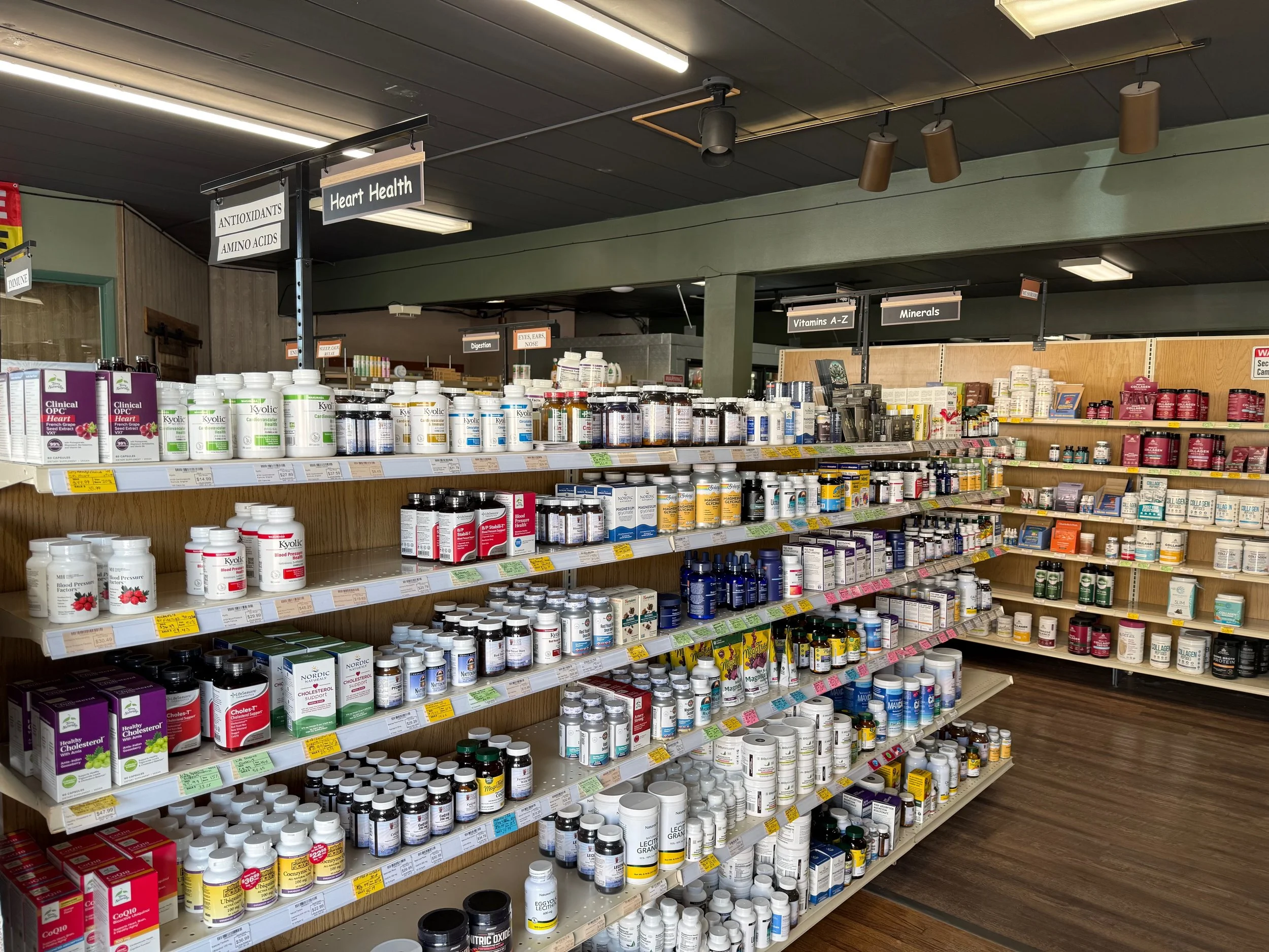 A store shelf stocked with various dietary supplements and vitamins, including bottles labeled 'Blood Pressure Factors,' 'Healthy Cholesterol,' and 'Cardiovascular Health.' Signs above the shelves indicate categories such as 'Antioxidants,' 'Amino Acids,' 'Heart Health,' 'Vitamins A-Z,' and 'Minerals.'