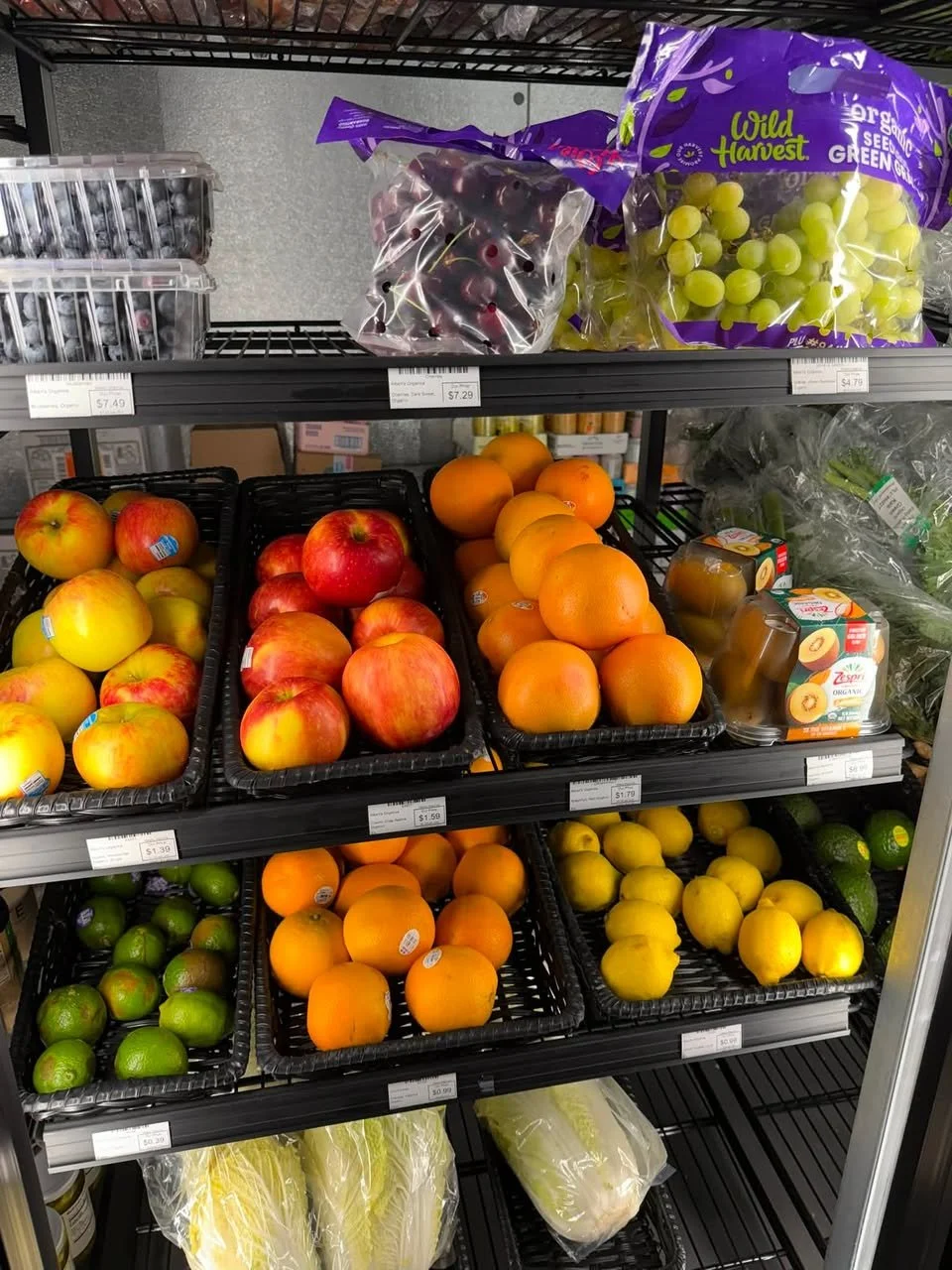 A grocery store shelf stocked with fresh fruits including apples, oranges, limes, grapes, and packaged lemon wedges.