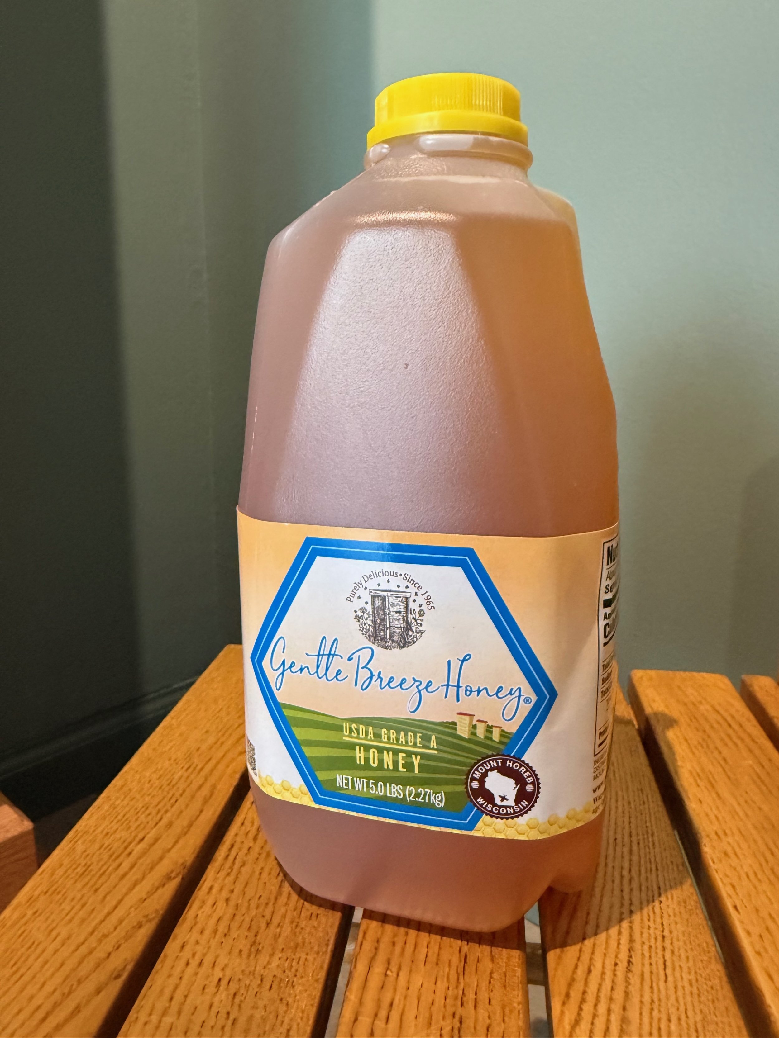 A plastic container of honey with a yellow cap, placed on a wooden surface. The label reads 'Gentle Breeze Honey,' indicating it is USDA grade A honey from Mount Horeb, Wisconsin.