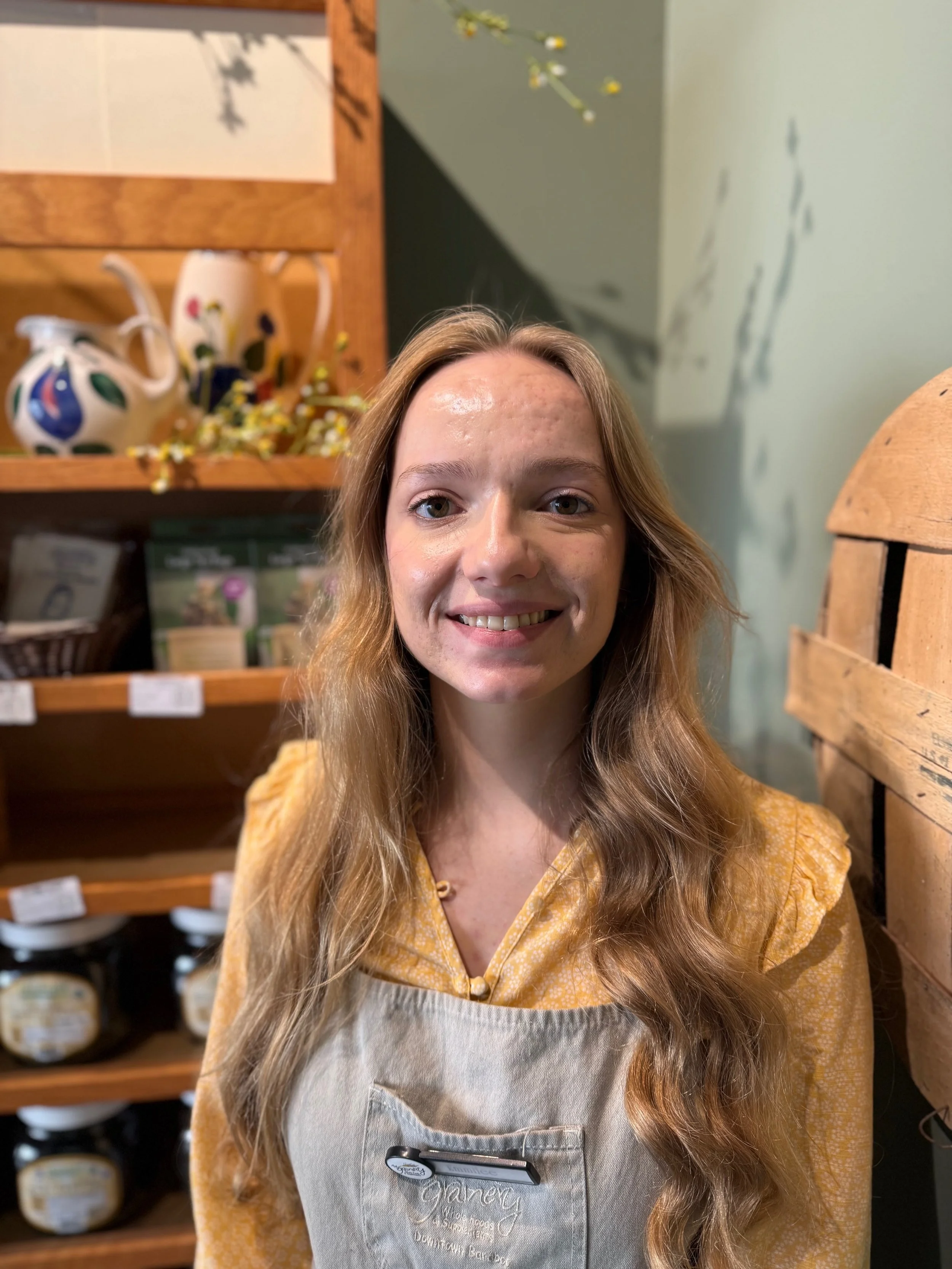 A young woman with long wavy blonde hair smiling at the camera inside a store with tiered wooden shelves in the background, wearing a yellow printed top and a light-colored apron with a name tag and a shopping list attached.