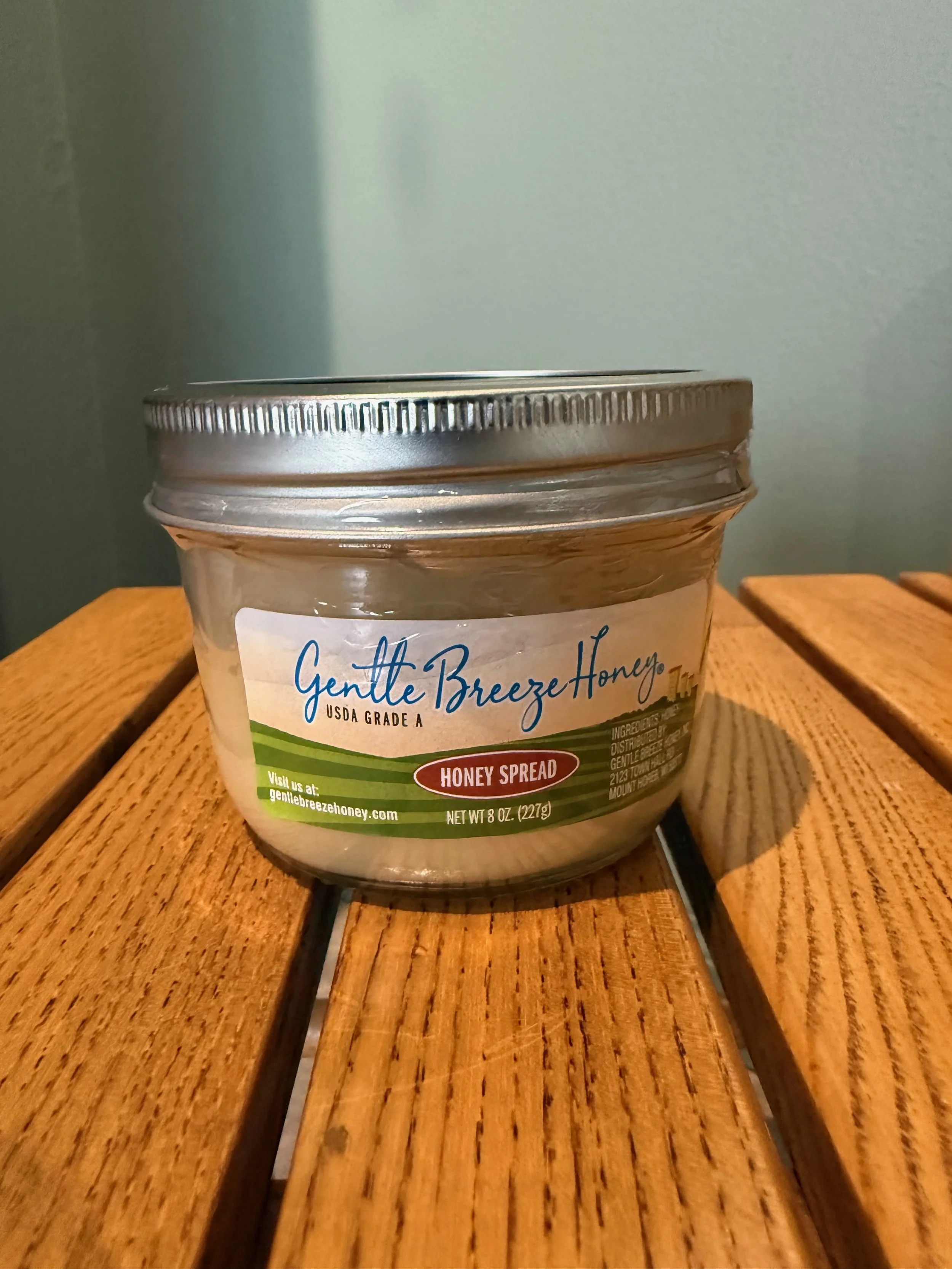 A jar of Gentle Breeze Honey, honey spread, placed on a wooden surface against a plain wall background.