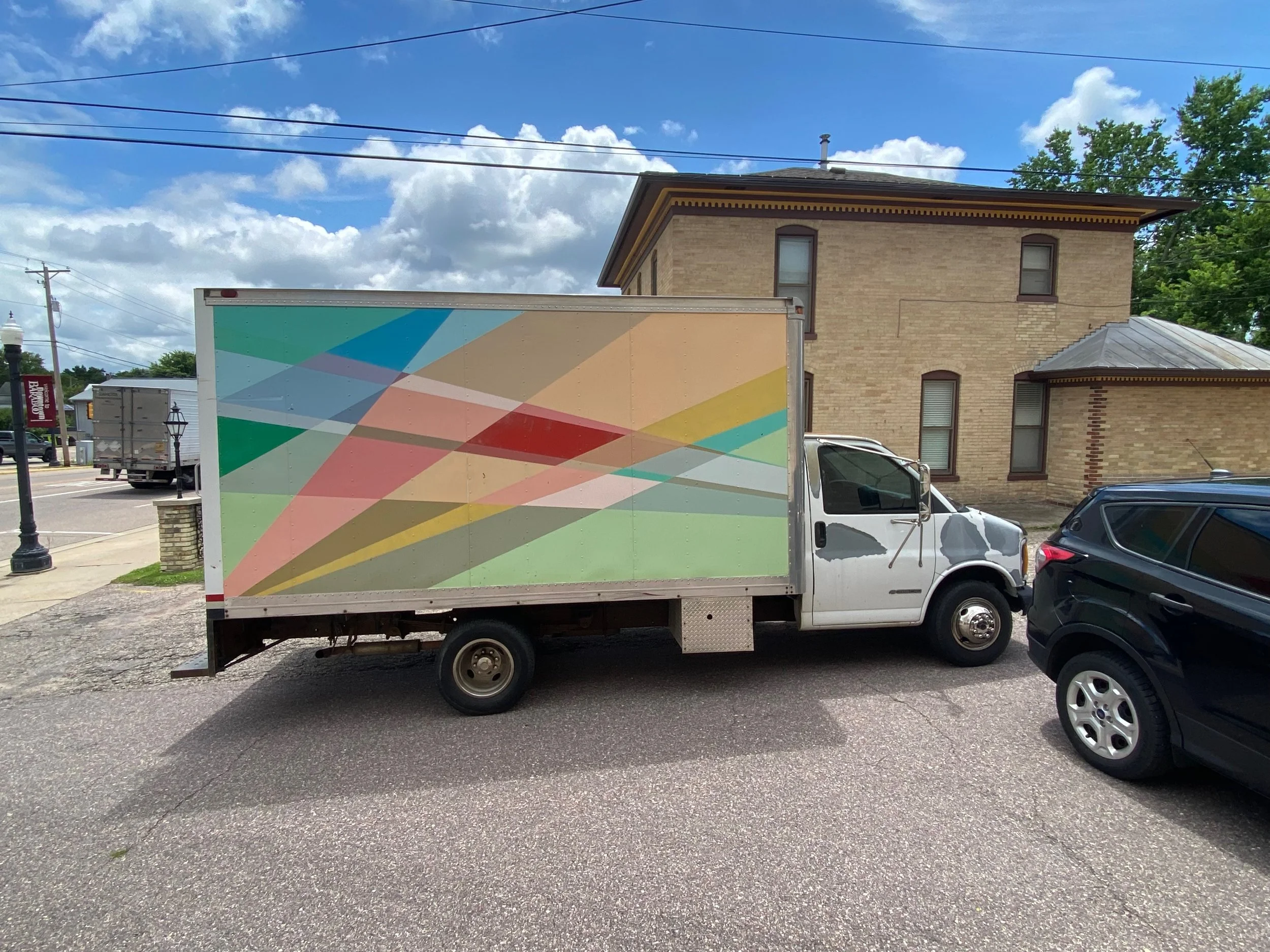 A white delivery truck with a colorful geometric pattern on its side parked on a street next to a black vehicle. In the background, there is a brick building with multiple windows, power lines, a lamppost, and a cloudy blue sky.
