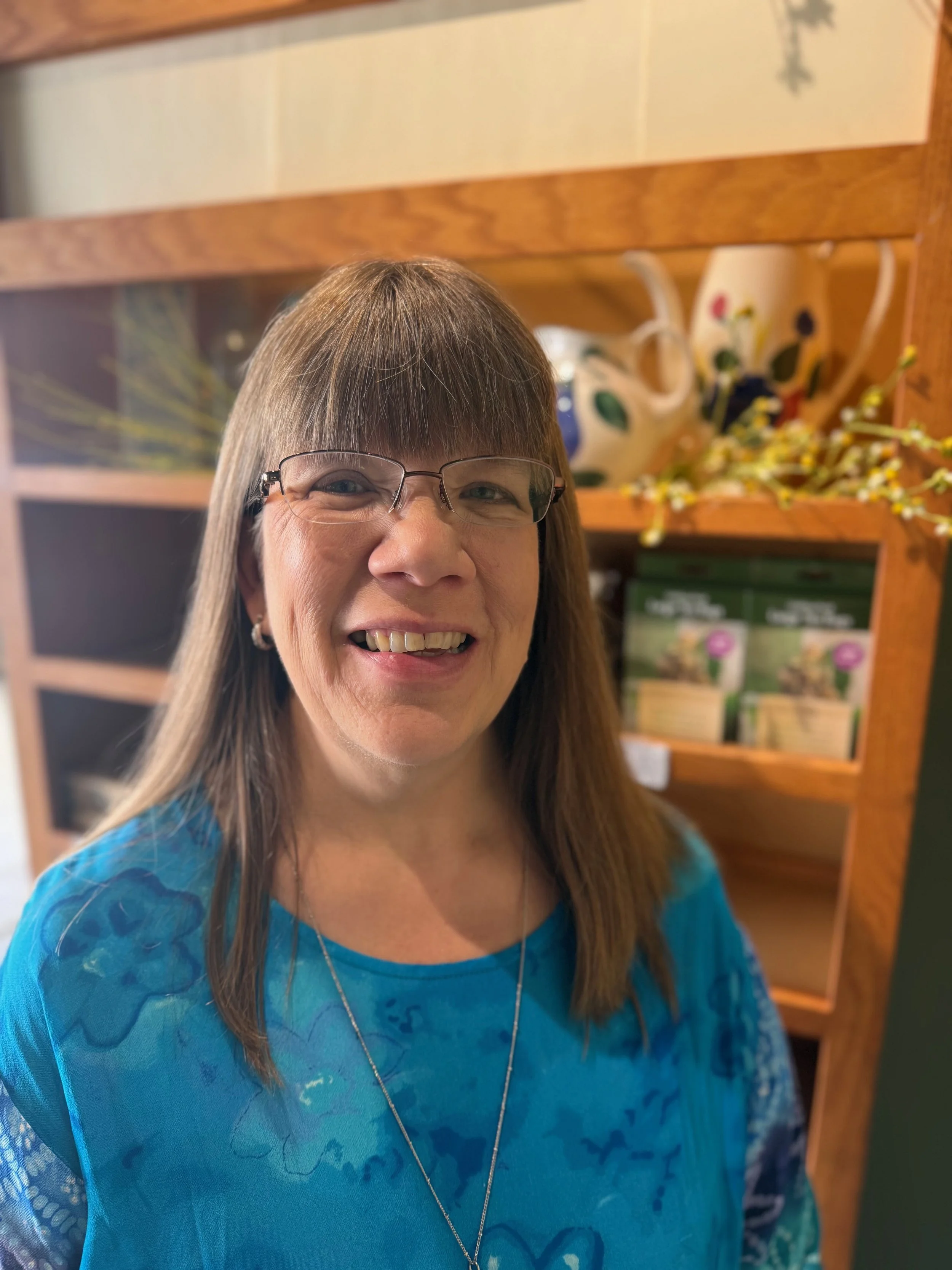A smiling middle-aged woman with glasses and shoulder-length brown hair, wearing a blue patterned top, standing in front of a wooden shelf with decorative items, including a ceramic teapot with colorful design and a box of garden seeds.