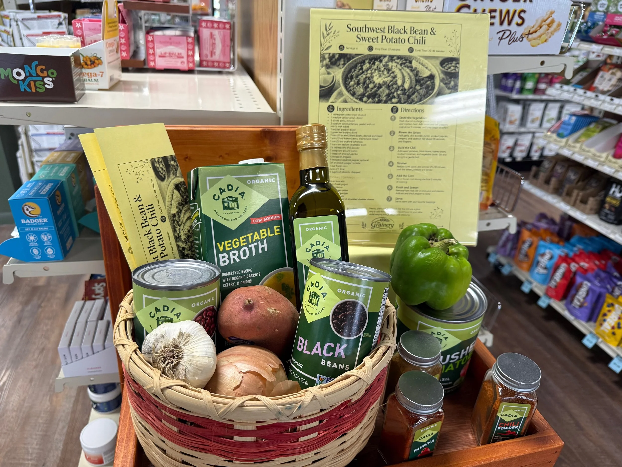 A display at a grocery store with cans of organic black beans, garlic bulbs, onions, and green bell peppers in a basket, along with a yellow recipe card for Southwest Black Bean & Sweet Potato Chili. The background includes various other food products and shelves.