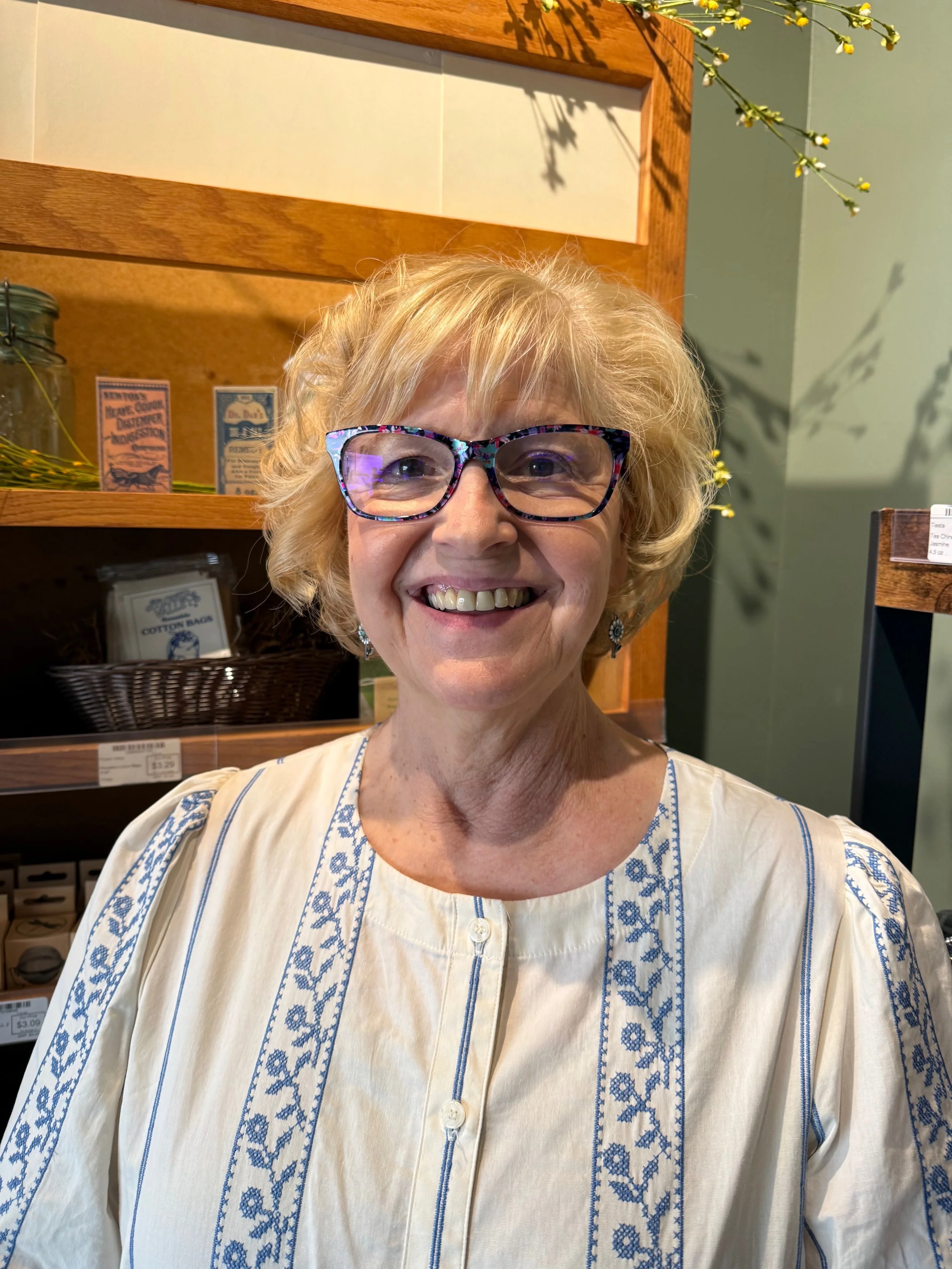 Close-up of a smiling woman with glasses and blonde hair, wearing a cream blouse with blue embroidery, inside a store with wooden shelves and products in the background.